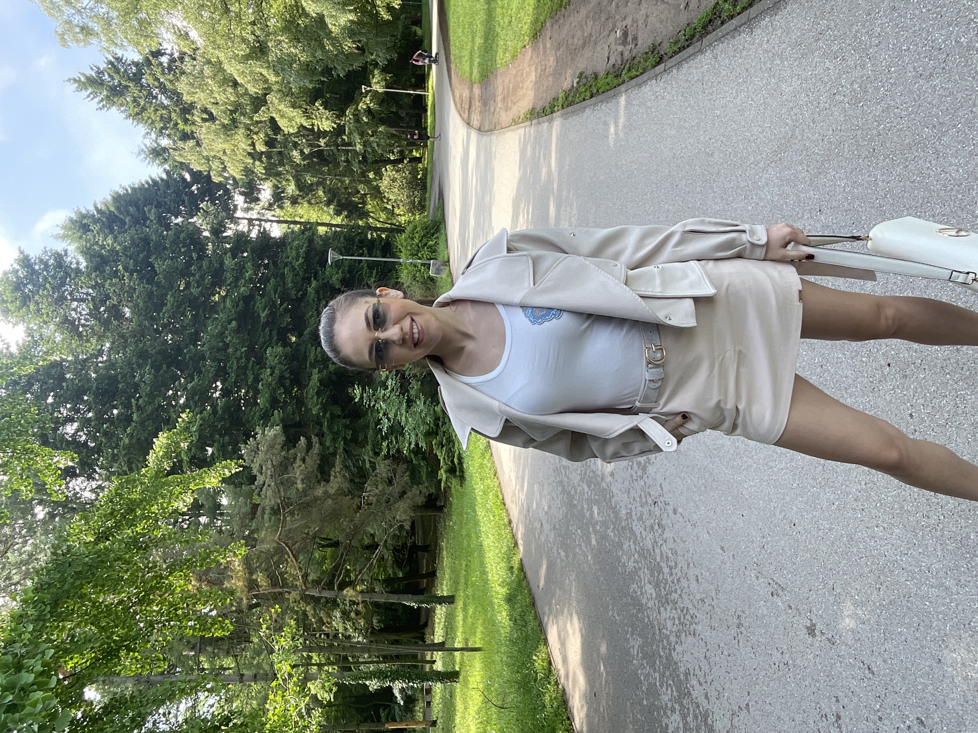 The image captures a smiling woman posing on a paved path in a sunny park in Sofia, Bulgaria. She is centrally positioned, looking towards the camera with sunglasses on and her hair pulled back. She wears a light-colored (beige/cream) jacket over a white tank top, which features a small blue and white emblem on the left chest area, and a matching light-colored skirt or shorts. A belt with a gold-colored buckle is visible, and she carries a white handbag with a gold-toned clasp.
The setting is a vibrant green park, with numerous mature trees (both coniferous and deciduous) lining the curving paved path. Lush grass covers the ground between the path and the tree line. In the distance, on the path, faint figures of other park-goers, including one on what appears to be a scooter, can be seen, suggesting a leisurely environment. The time of day is clearly daytime, with bright sunlight filtering through the tree canopy and casting shadows on the path. The weather appears clear and pleasant. The woman's expression conveys a cheerful and relaxed mood. No legible text is visible in the image.