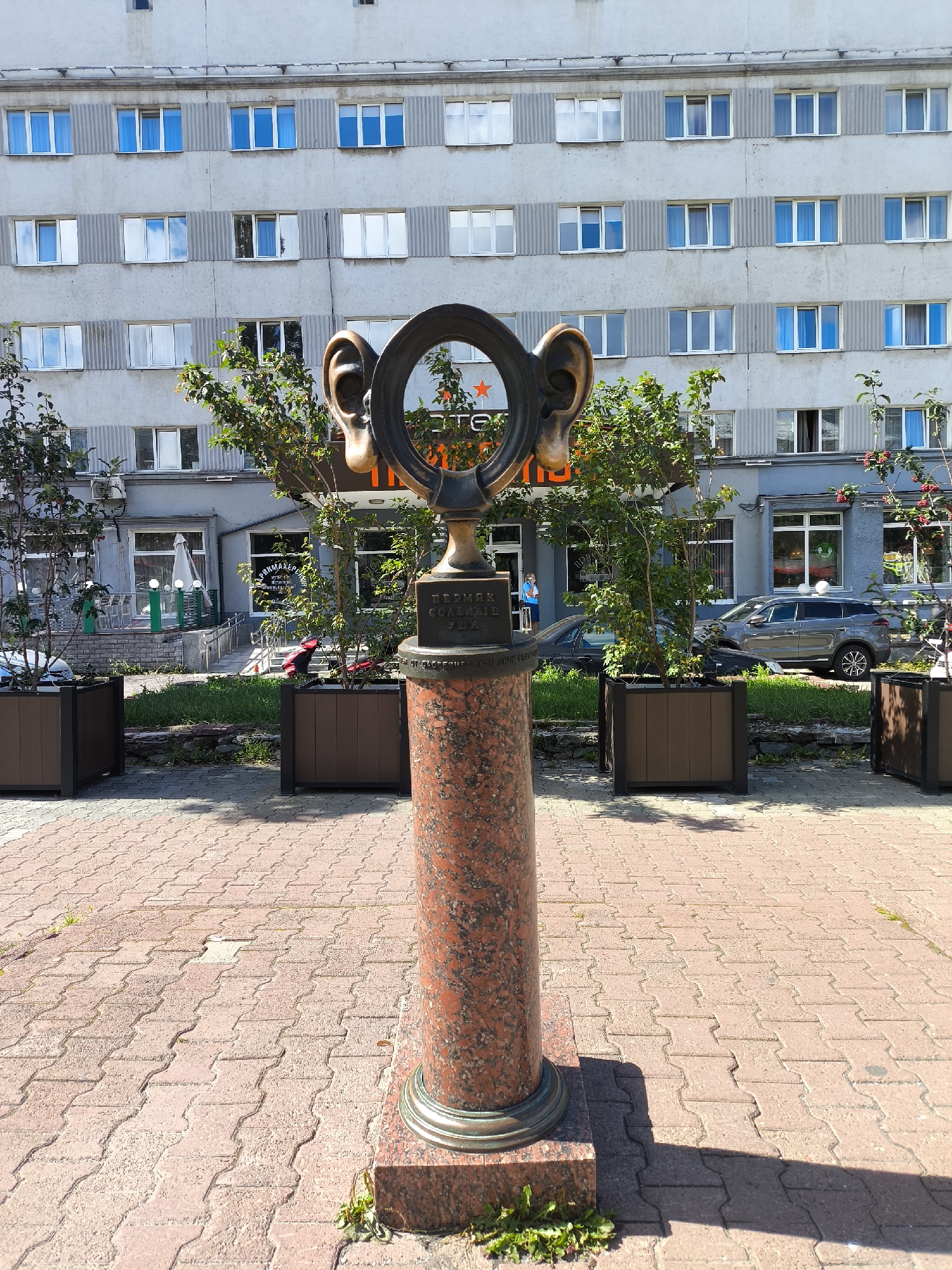 The image shows a sculpture of a large ear with a ring in the middle, standing on a red marble pedestal. The sculpture is located on a sidewalk paved with reddish-brown interlocking bricks, in front of a light gray, multi-story building with many windows. There are green trees planted in brown rectangular planters on either side of the sculpture. In the background, businesses with storefronts are visible. The sculpture appears to be a public art installation. The inscription on the pedestal is partially visible and reads "ПЕРВАЯ... КОМПАНИЯ", which translates to "First Company". This sculpture is known as the "Ear of Gastell" and is located on Perm-1 train station square in Perm, Russia.