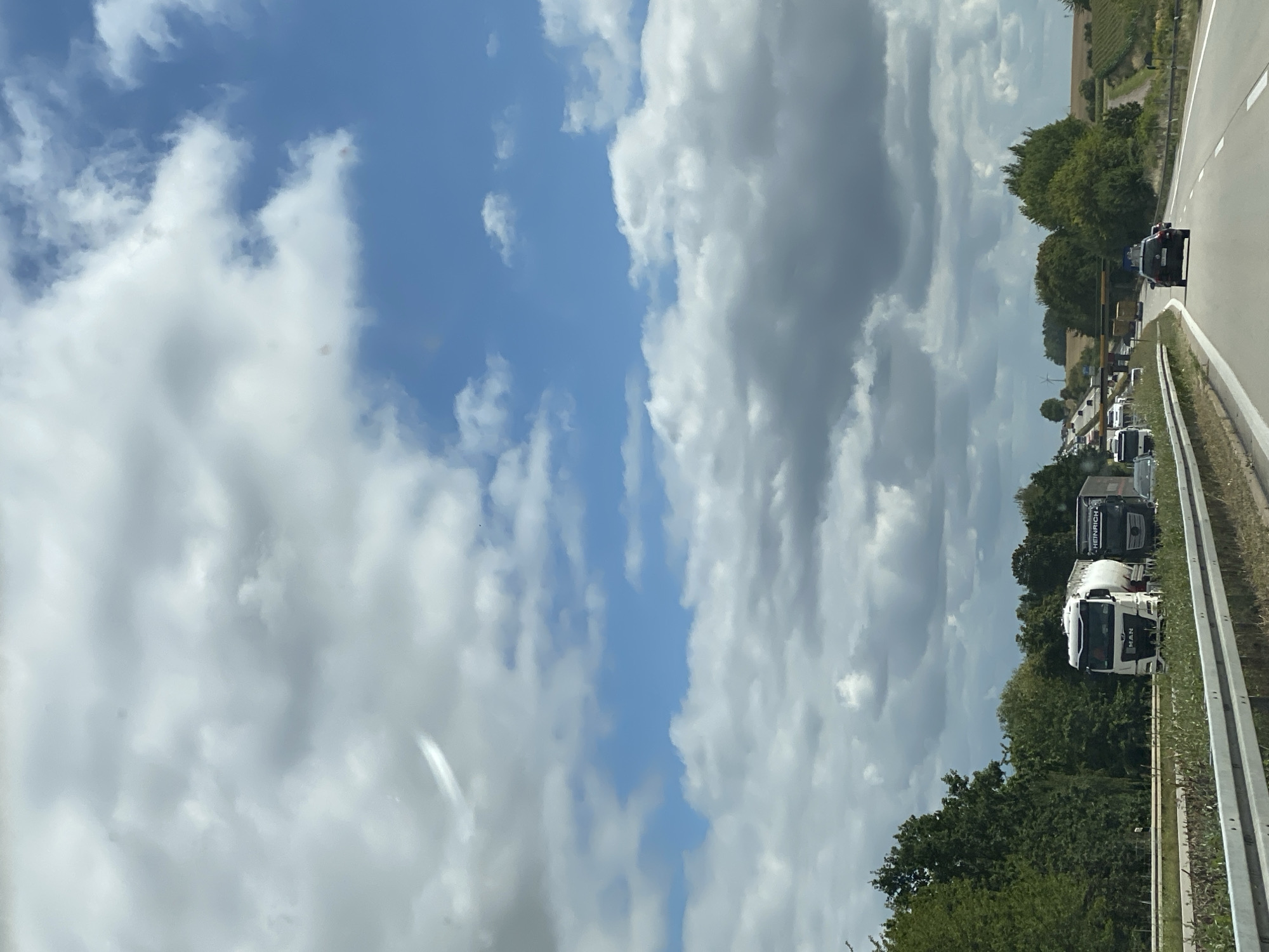 The image shows a highway scene with several trucks and cars. The sky is partly cloudy with blue patches. On the left side of the highway, there is a dense line of trees. On the right side, there are agricultural fields. In the foreground, there is a guardrail and vegetation. Several large trucks, including a white tanker truck and a black truck with "HEINRICH" written on it, are visible on the highway. Cars are also present, with one black car in the right lane ahead of the viewer. The scene appears to be a traffic jam or slow-moving traffic, as evidenced by the congestion of vehicles in the distance. The overall impression is a daytime highway scene in a rural or semi-rural area, possibly in Germany given the reference to Döbeln.