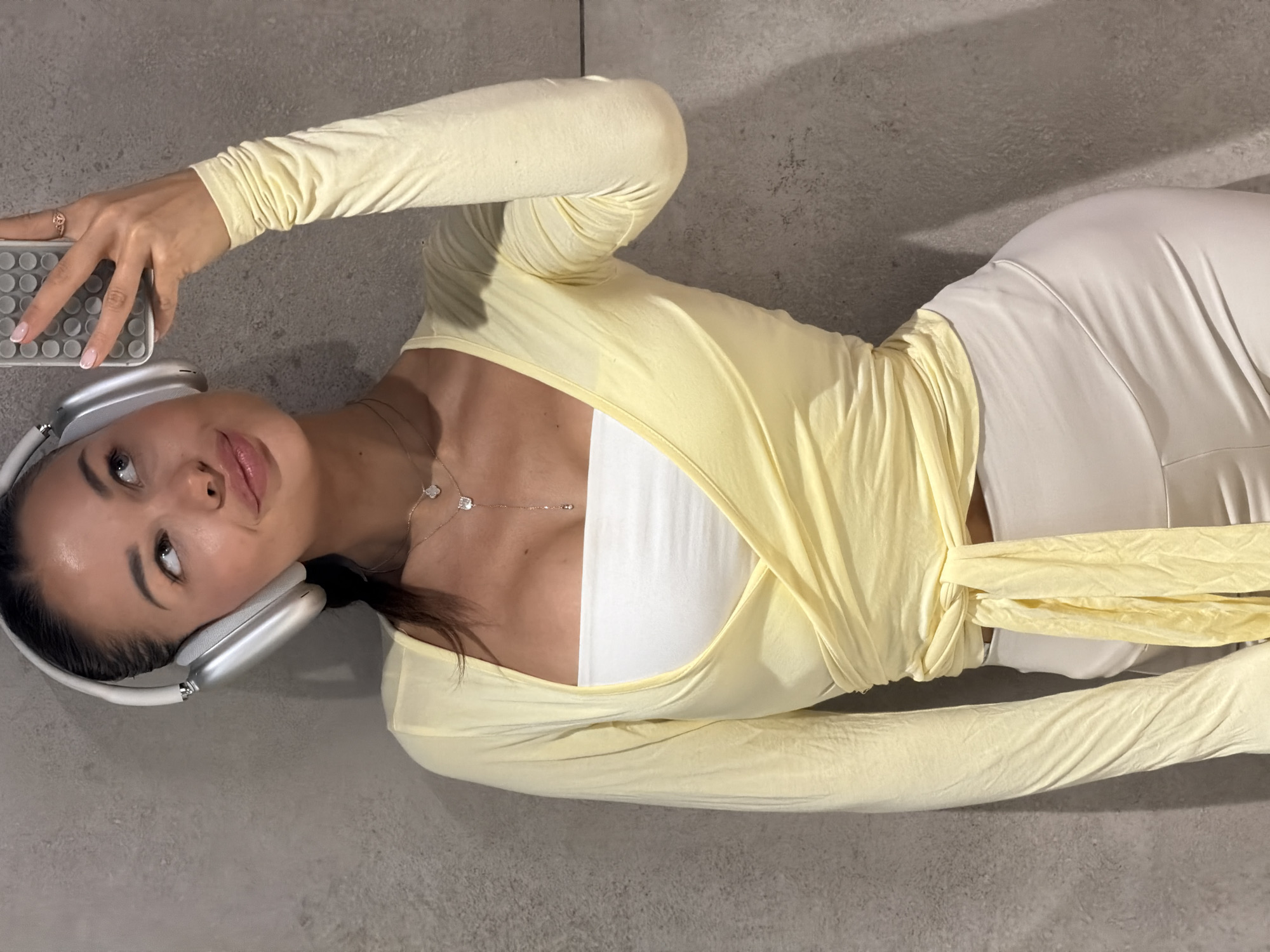 The image features a young woman lying on her back on a light gray, possibly tiled or concrete, floor. She is positioned diagonally across the frame, looking slightly upwards and to the right with a serene expression and a subtle smile. She wears a light yellow long-sleeved top that wraps around her torso, layered over a white crop top. Her lower body is clad in light-colored, possibly white or cream, form-fitting bottoms.
On her head, she wears silver over-ear headphones, resembling Apple AirPods Max. Her left hand, with a ring on her ring finger and manicured nails, holds a smartphone encased in a distinctive gray, textured silicone cover, featuring a pattern of raised circular bumps. A delicate silver chain necklace with two small pendants, one clover-shaped and one square, adorns her neck. Her dark hair is pulled back, and she has natural-looking makeup. The lighting is even, suggesting an indoor setting without harsh direct sunlight.
The activity depicted is a relaxed pose, possibly a selfie or a casual moment while listening to music. There is no visible text in the image. The setting is indoors, with the floor being the most prominent background element. The overall impression is one of calm and modernity. The context provided indicates the location is Moscow, Russia.