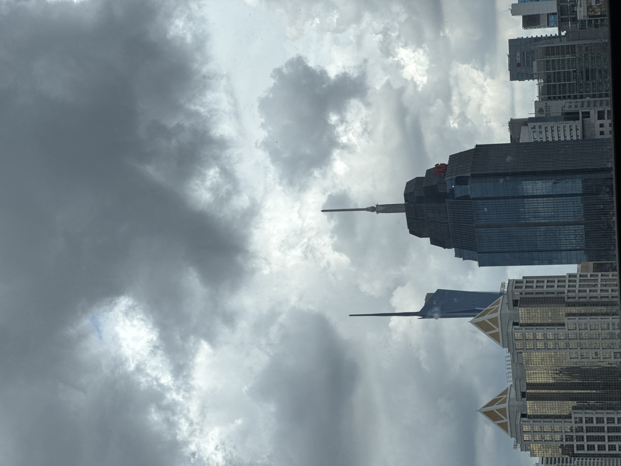 This image captures a portion of the Kuala Lumpur skyline under a cloudy sky. Prominent skyscrapers dominate the frame, with a dark, textured building featuring a red logo on its upper section centrally positioned. To its left, a lighter-colored building with geometric, pyramid-like roofs and reflective gold windows is visible. Further to the left and slightly behind, a tall, slender blue skyscraper tapers to a sharp point. Several other buildings of varying heights and architectural styles are scattered across the background, some with visible signage. The sky is filled with heavy, grey clouds, with a few brighter patches suggesting diffused sunlight. The overall impression is of a dense urban environment on a potentially overcast day.