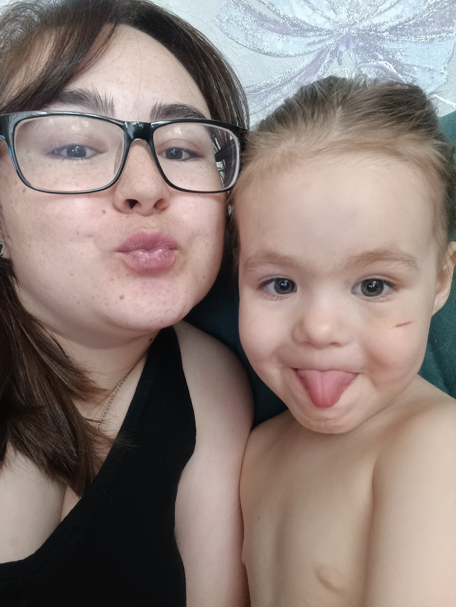A selfie depicts a woman with glasses and a young child. The woman has dark hair, fair skin with freckles, and wears black framed glasses. She is making a kissing face toward the camera and wears a black tank top with a silver necklace. The child, next to her, has light hair pulled back, and is sticking her tongue out, with a scratch on her right cheek. The child is shirtless. The background is a light wall with a floral pattern. The location is Noviy, Russia.