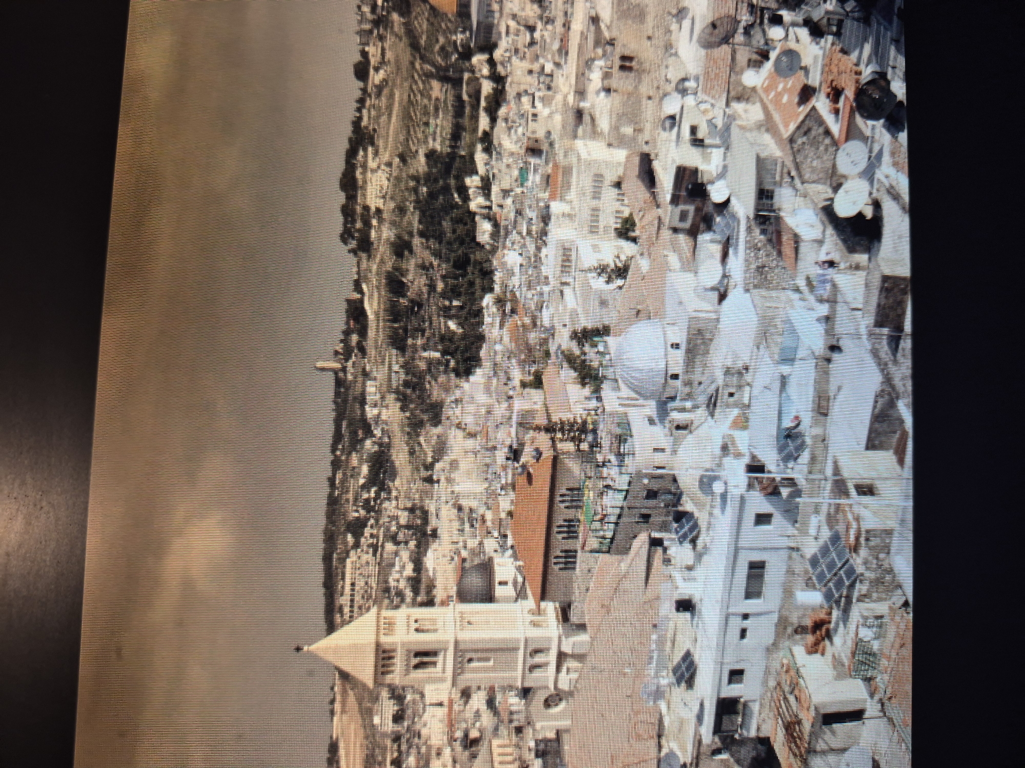 The image is a sepia-toned aerial view of a dense cityscape. A hill with vegetation is on the left. The buildings are densely packed together with various roof types including flat, tiled, and domed structures. The buildings range in height from one to several stories. Satellite dishes can be seen on some rooftops. In the lower center is a building with a tall, pyramid-shaped spire. The sky is overcast. The location is not Yerevan, Armenia. The city is Jerusalem.