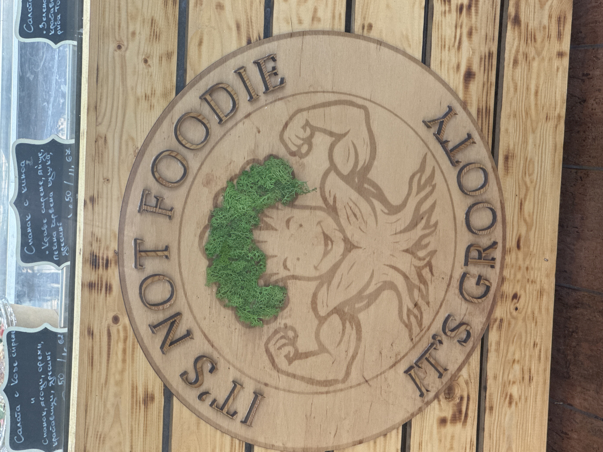 The image shows a circular wooden sign with carved text and a cartoonish illustration of a muscular tree-like character with green moss for hair. The text on the sign reads "IT'S NOT FOODIE" and "IT'S GROOTY". The sign is mounted on a wooden plank wall. Above the sign, there is a display of food items with handwritten labels in Bulgarian. The labels mention salads and ingredients. The context suggests this is a sign for a food establishment or a stall at a market in Sofia, Bulgaria, likely selling healthy or plant-based food.