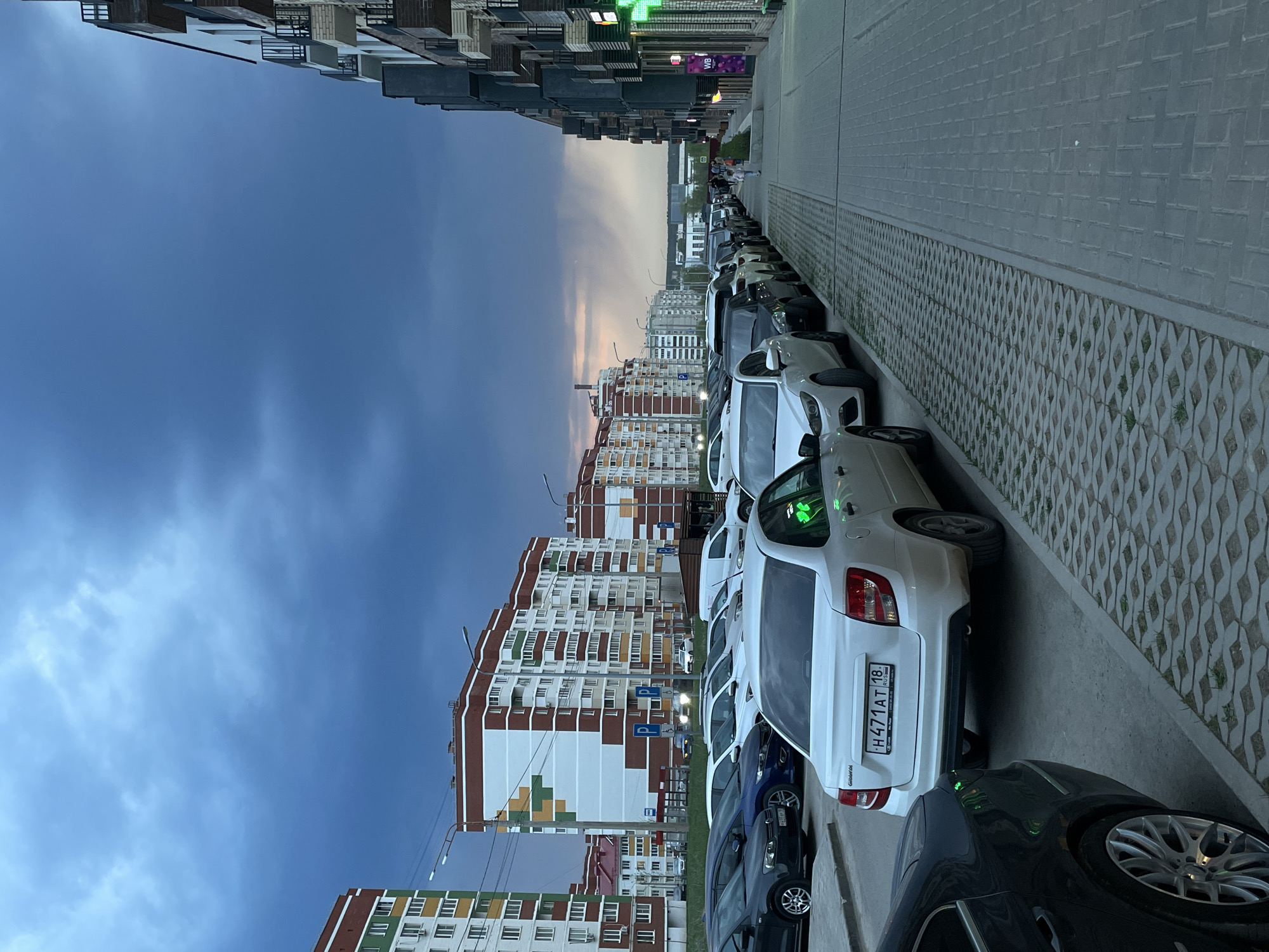 A street scene in Izhevsk, Russia, shows a row of parked cars along a sidewalk. The license plate on a white car reads "H471AT 18," indicating the car is registered in Udmurtia (region code 18). Buildings line both sides of the street, including tall apartment complexes with distinctive architectural features and color schemes. The sky is blue with hints of sunset. Pedestrians are visible in the distance.