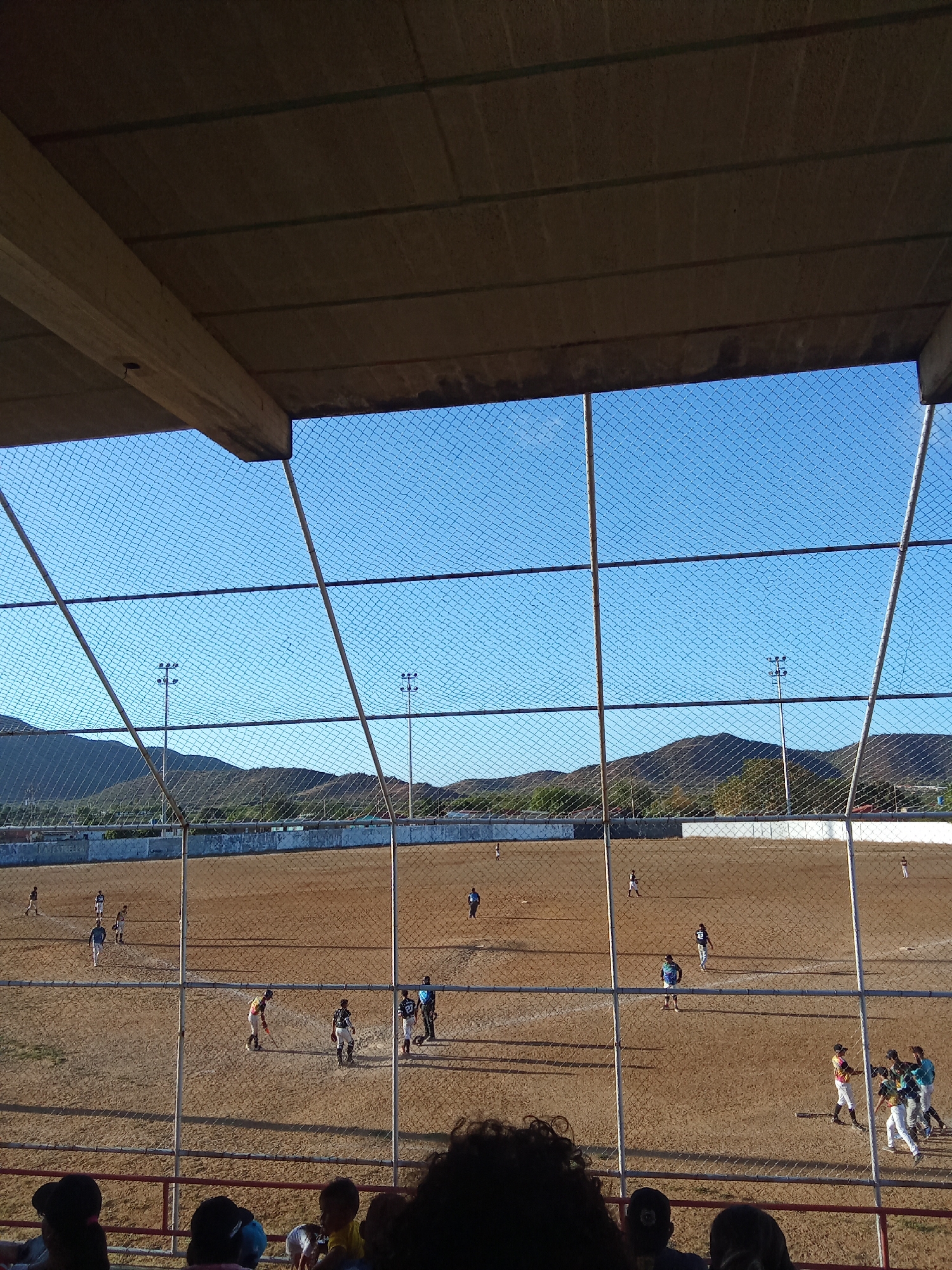 The image shows a baseball game in progress on a dirt field in Juan Griego, Venezuela. The view is from under the roof of a spectator stand, looking through a chain-link fence. Several players in uniform are on the field, some in groups. There are mountains in the background under a clear blue sky. Stadium lighting poles are visible along the periphery of the field. Spectators are seated in the foreground, in front of the fence.