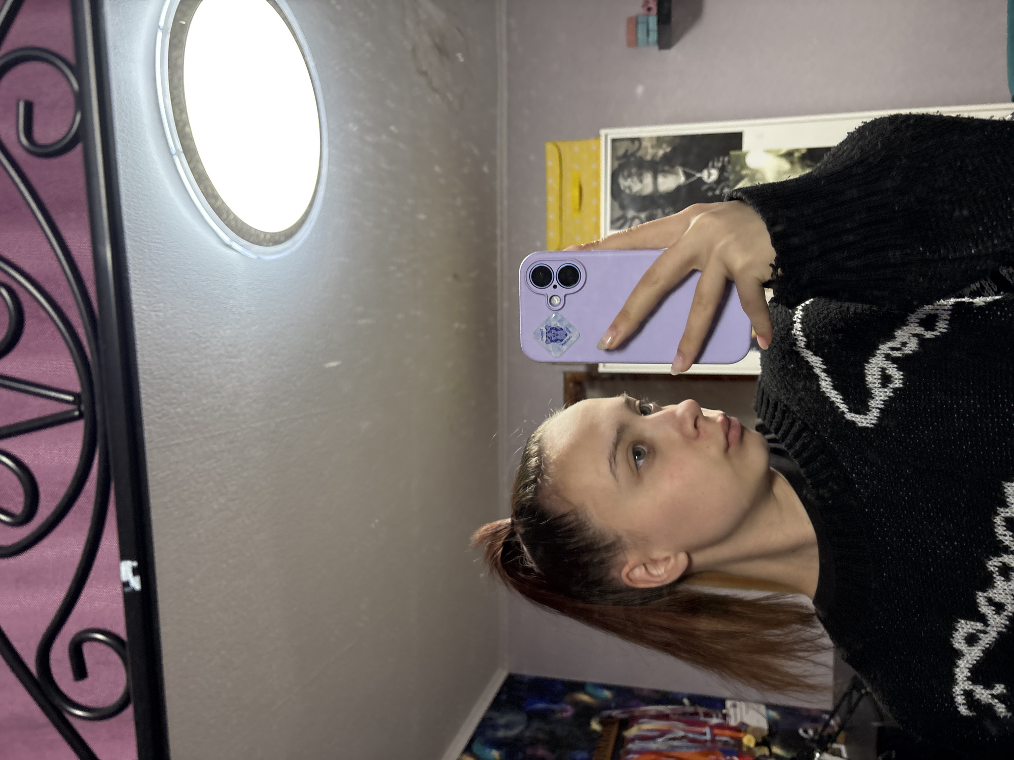 A young woman is taking a selfie in a room. She is wearing a black sweater with white patterns. She is holding a purple phone. Behind her are a circular light fixture, a metal decorative piece, and framed artwork. The image was taken in Прикамский, Russia.