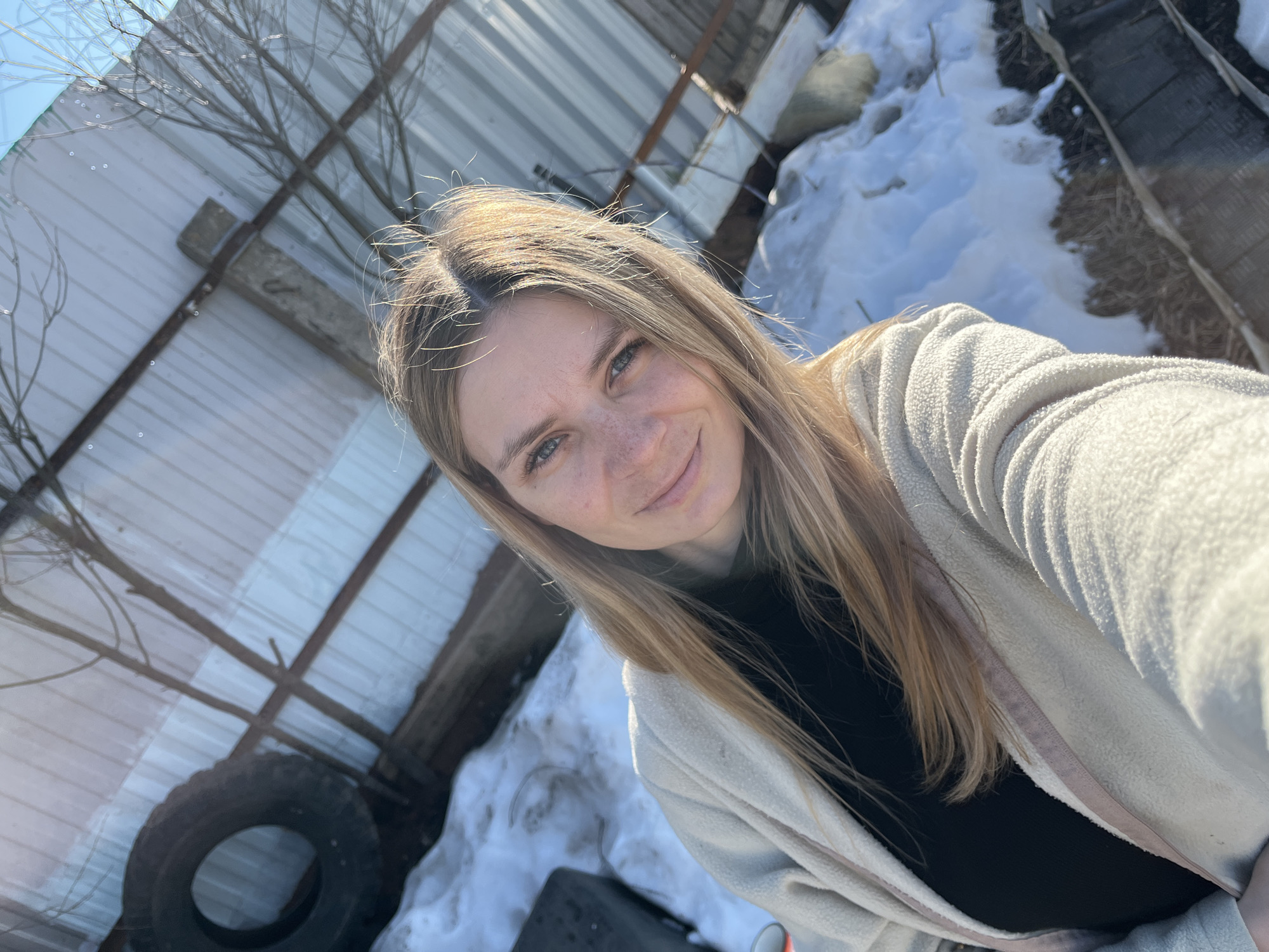 A selfie of a woman with light skin, freckles, and light brown hair, wearing a light-colored cardigan over a black top. She is smiling slightly at the camera.
The background is a somewhat cluttered outdoor scene. In the top left is a corrugated metal fence or roof structure with wooden support beams and bare tree branches. Below this, there is a black tire. To the right of the woman is a snow pile, and a dark object is partially visible at the bottom of the frame.
The location may be near Chaikovsky, Russia, as specified.