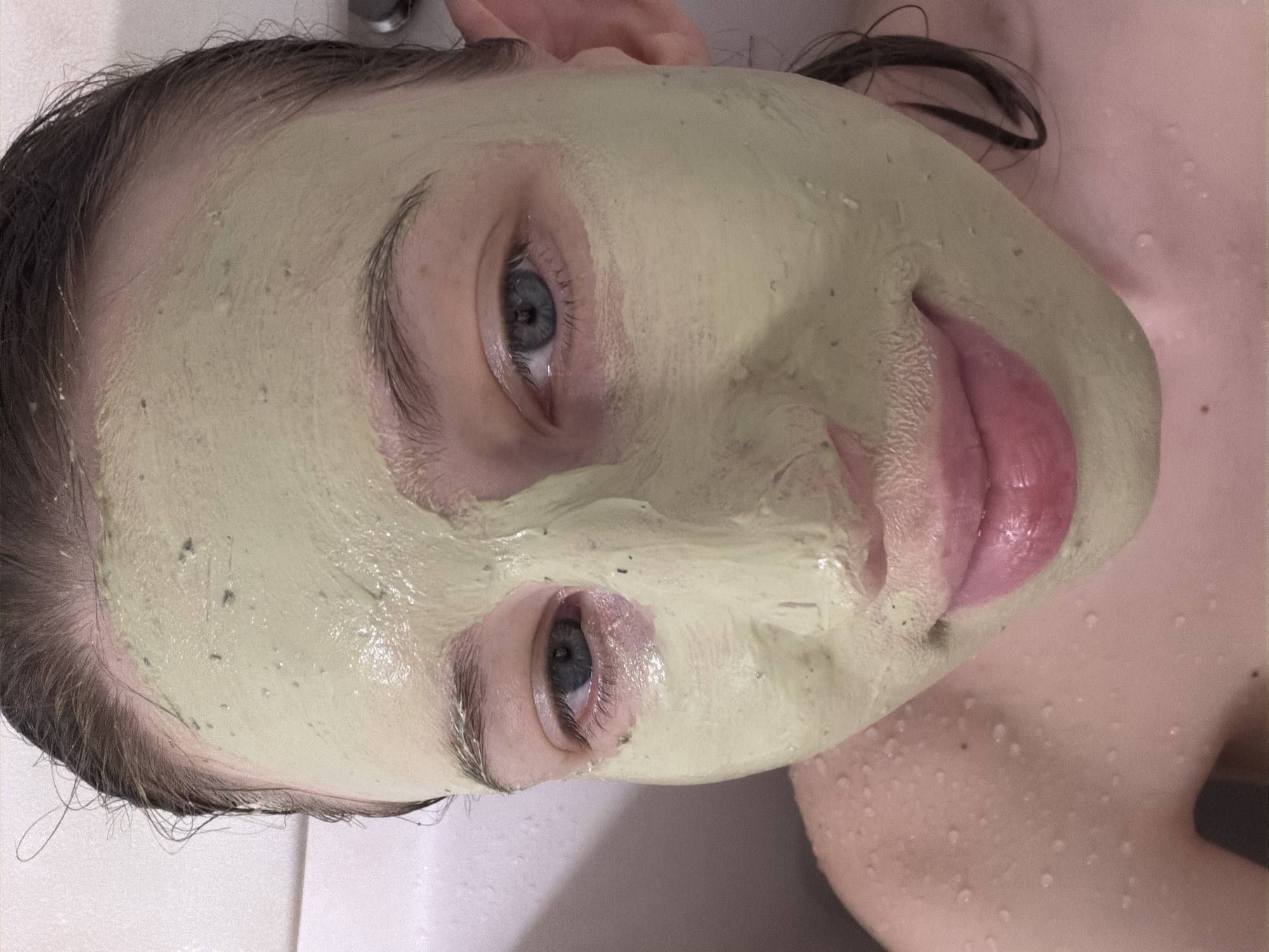 A close-up, waist-up shot shows a person with a pale complexion, wearing a light green facial mask and a pink lip balm. The mask covers the entire face, obscuring the nose, and extends to the hairline. The subject has gray-blue eyes and dark brown hair, which appears wet and is pulled back from the face. Water droplets are visible on the subject's shoulders and chest, suggesting they are in a bathroom or shower. The background is a plain, light-colored wall. The lighting is soft and diffused, creating a gentle illumination of the subject. The image is tightly cropped, focusing on the face and upper body. The location in Dongducheon-si, South Korea is not discernible from the image itself.