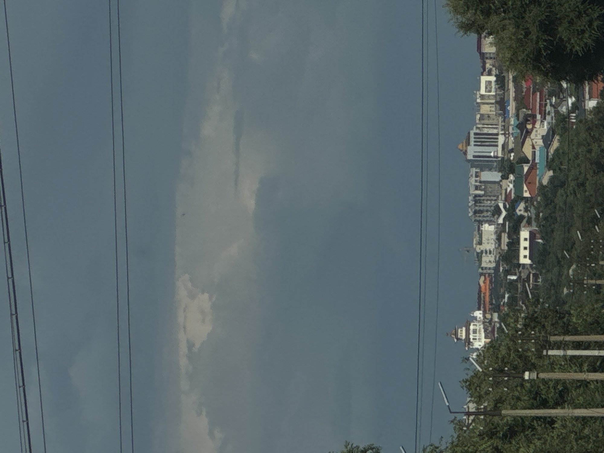 This media file depicts a distant view of a city skyline in Dorodny, Russia. The image is oriented vertically, with the cityscape appearing on the right side against a hazy, blue sky. Several power lines run diagonally across the left portion of the frame, creating a strong linear element.
The cityscape is characterized by a collection of buildings, including what appear to be modern multi-story structures mixed with older, lower-rise buildings with pitched roofs. One prominent building in the mid-ground has a distinctive pyramid-like roof. The architecture suggests a developing urban area. Dense green foliage, likely trees, borders the cityscape on the right, obscuring some of the lower portions of the buildings and creating a natural foreground element.
The lighting suggests it is daytime, possibly late afternoon or early morning, given the soft light and muted colors. The sky is mostly overcast with some patches of white clouds, indicating fair to partly cloudy weather. There are no visible people or specific activities depicted; the focus is on the static view of the urban landscape and the surrounding natural elements. No text is discernible within the image.