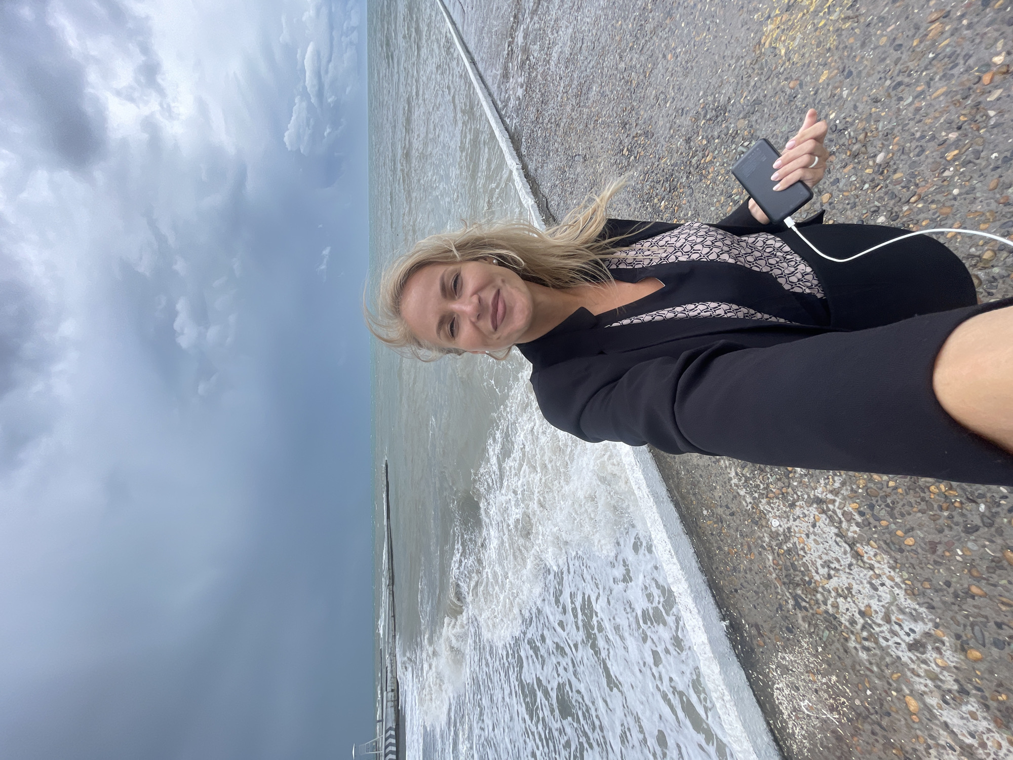 A woman is taking a selfie at the coastline with crashing waves and a cloudy sky. She is holding a phone and a charger. The location is not identifiable as Sochi, Russia.