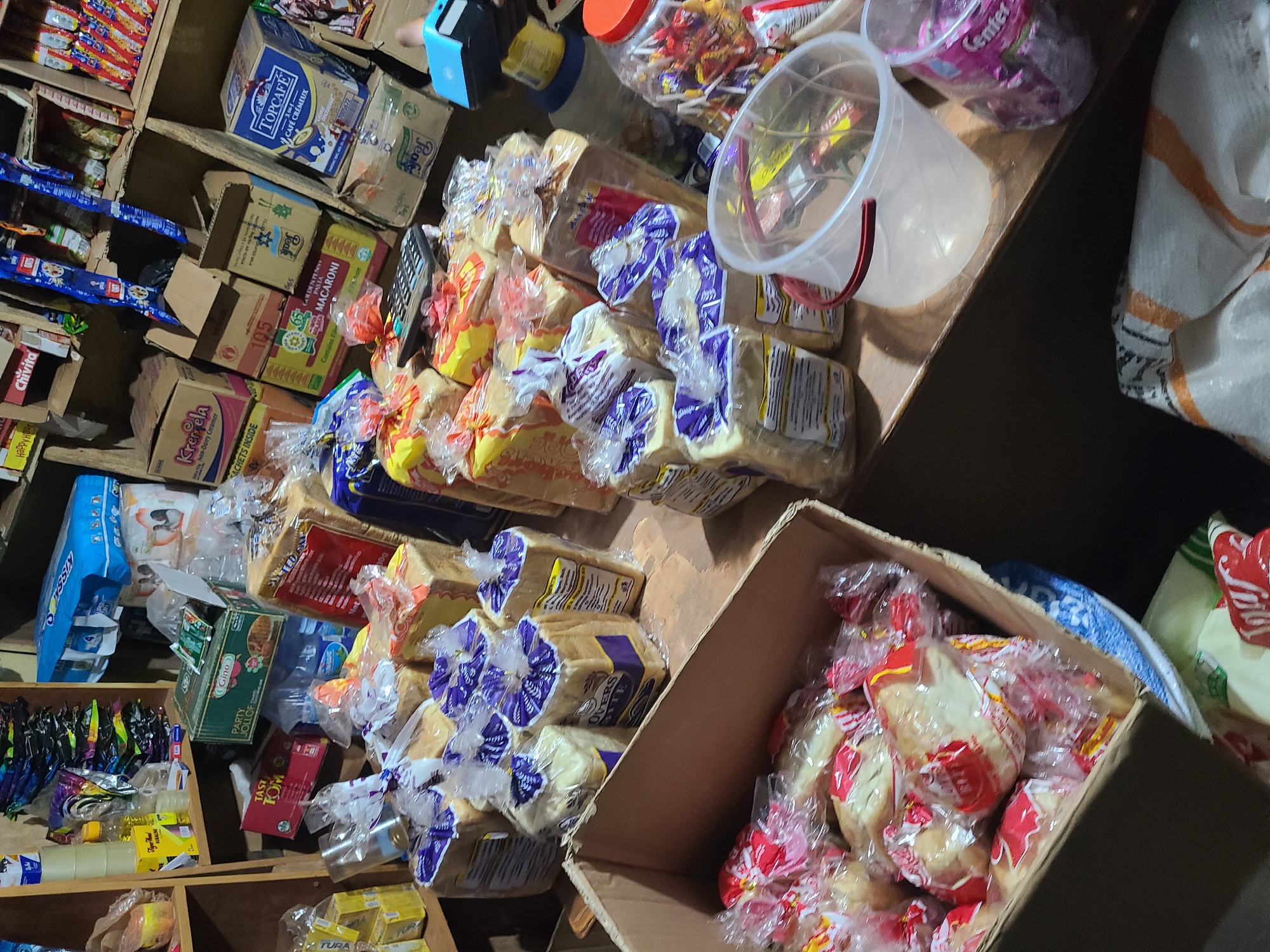 The image depicts a cluttered store interior, likely a small shop or market stall, located in Abuja, Nigeria. The shelves are overflowing with a diverse array of products, primarily food items and packaged goods. Prominently displayed are numerous loaves of bread, mostly wrapped in clear plastic with colorful ties, some featuring purple and white labels. Other visible items include boxes of "TOPCAFÉ" coffee, "Gino PARTY JOLLOF" seasoning, and various other packaged foods with text in different languages, suggesting a local and possibly imported selection. Several clear plastic buckets and containers are also scattered among the products, one appearing to hold an assortment of candies or small packaged sweets. A calculator rests on a shelf near some of the bread, hinting at ongoing transactions. The lighting appears to be artificial, casting shadows and highlighting the density of the merchandise. The overall impression is one of a busy, well-stocked retail space. There are no people visible in the frame.