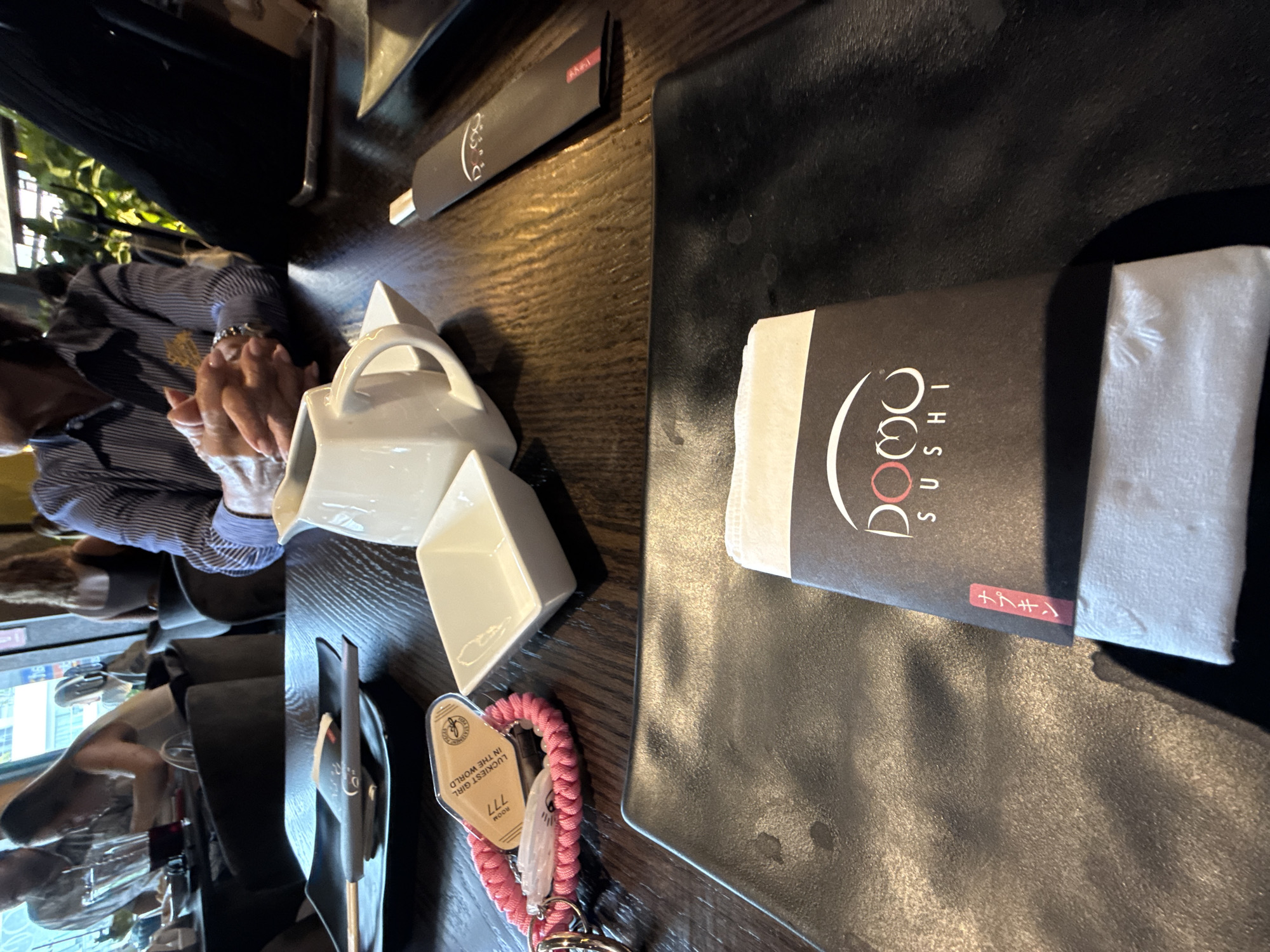 The image depicts a restaurant table setting, possibly at Domo Sushi, evident from the napkin packaging. A white ceramic teapot and a small square bowl are present. Cutlery in a black holder and chopsticks are visible. A pink braided bracelet with a "Luckiest Girl in the World" tag and "ROOM 777" are present. An individual is seated at the table with their hands clasped. The location appears to be San Bernardino Tlaxcalancingo, Mexico based on the context.