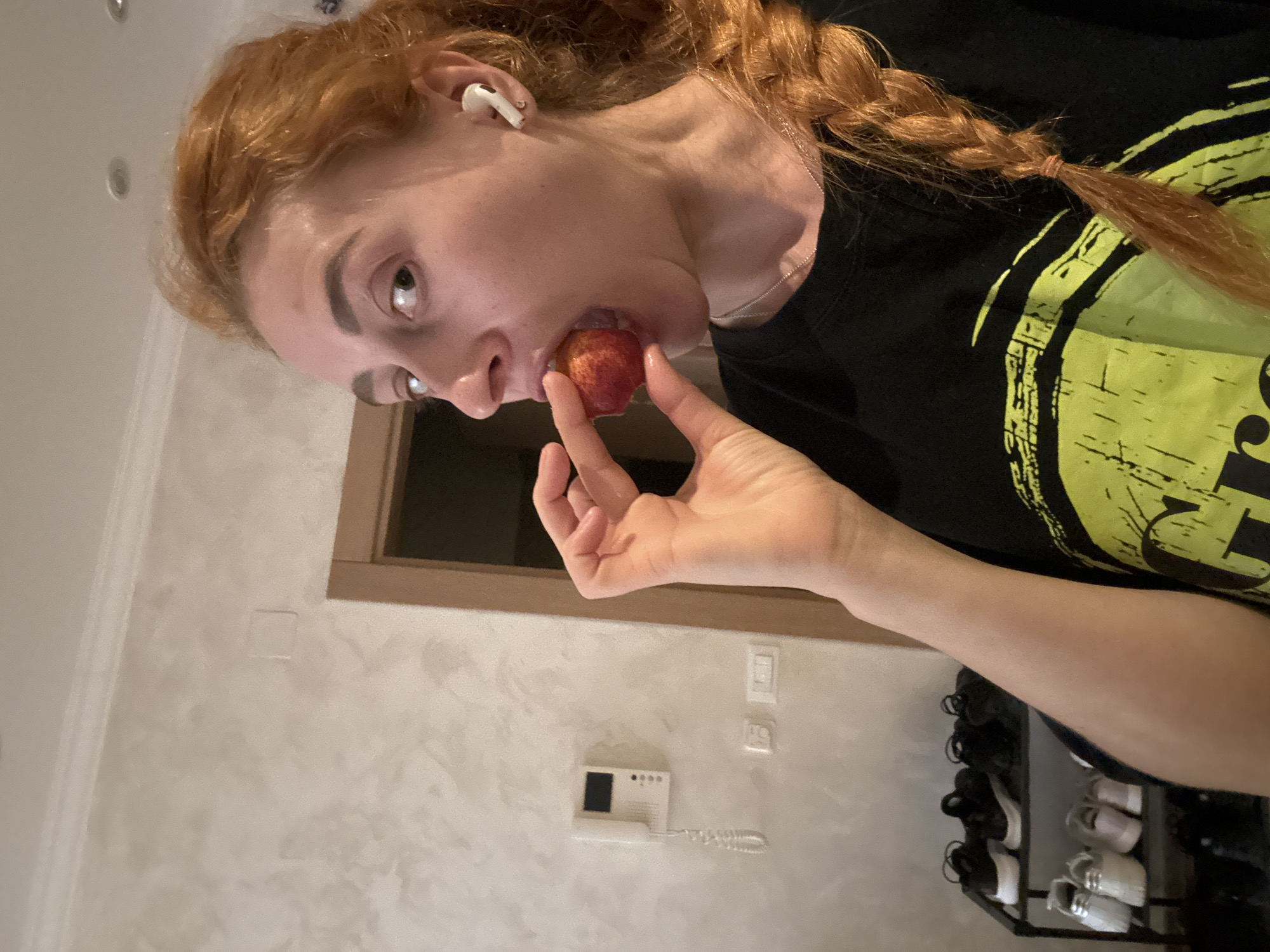 A reddish-haired woman with a braid and wearing an earpiece is eating a small red fruit. She is wearing a black t-shirt with a yellow design. She is standing in a room with a textured beige wall, a light wood-trimmed door, a wall-mounted intercom, a light switch, and a shoe rack with shoes on it. There is no information about the city of Budva, Montenegro in the image.