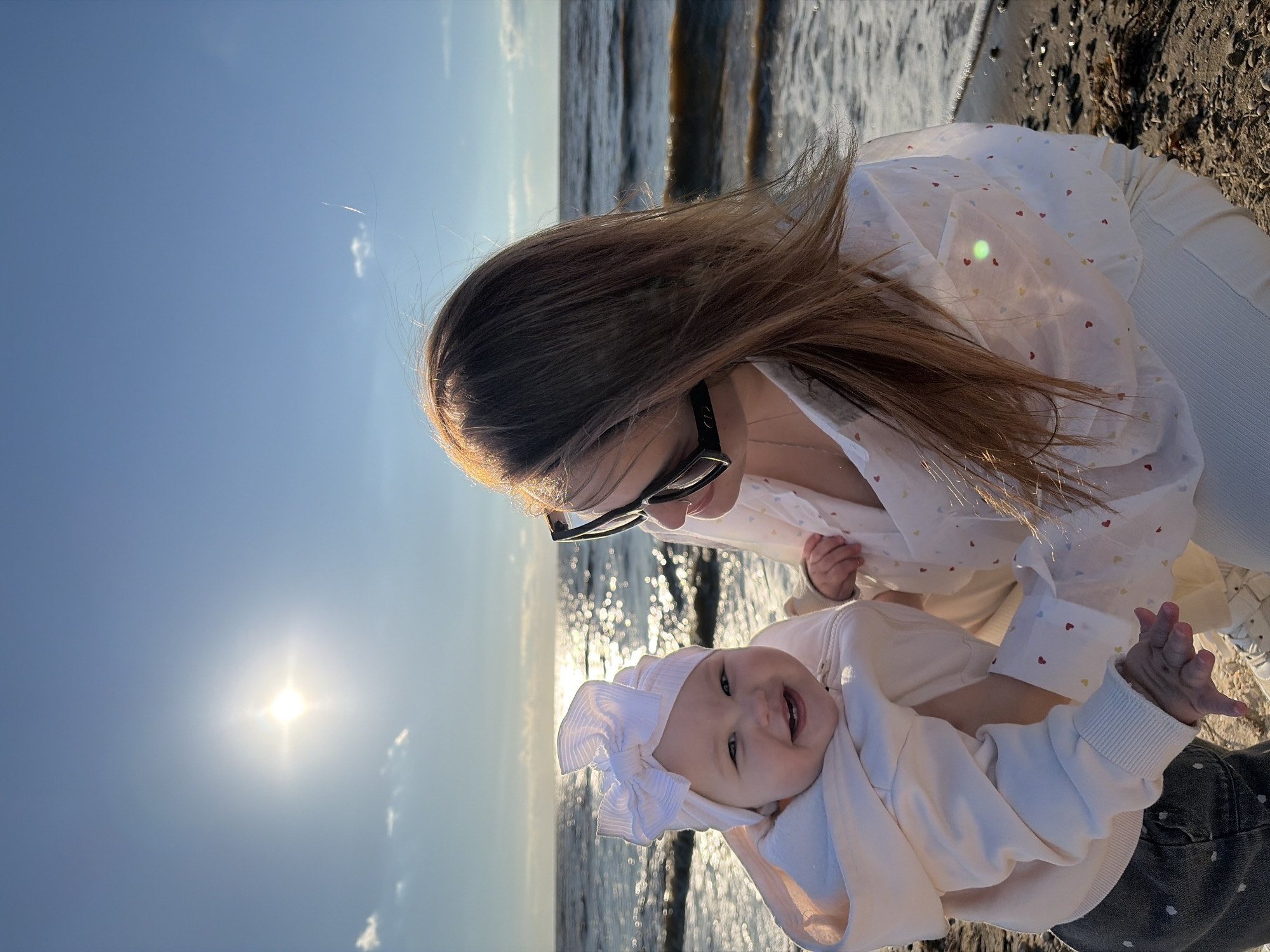 In Port-Katon, Russia, a woman wearing sunglasses and a white shirt with heart patterns holds a baby on a beach. The baby, wearing a white bow and a cream-colored hoodie, smiles at the camera. The sun shines brightly in the blue sky, reflecting on the water, and waves lap against the shore. The beach is covered in sand and small stones.