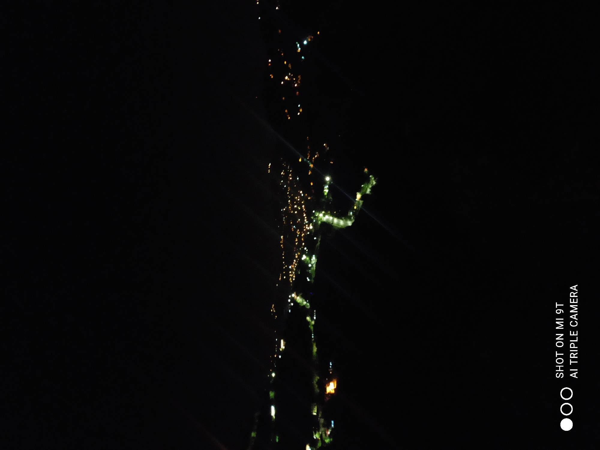The image is a nighttime aerial shot of a cityscape, likely from an elevated viewpoint overlooking the city of Akçaabat, Turkey. The darkness of the night sky is punctuated by numerous clusters of yellow and white lights, indicating buildings, streets, and possibly streetlights. Some lights appear to be concentrated in the distance, suggesting a central urban area, while others are more spread out. There is a visible band of lights that follows a curved path, possibly representing a road or a river winding through the city. The overall impression is a sprawling urban landscape illuminated at night. The bottom of the image contains a watermark with white text reading "SHOT ON MI 9T AI TRIPLE CAMERA".