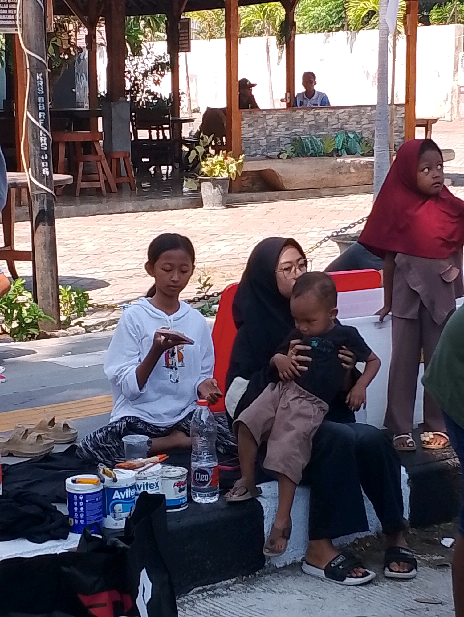 The image shows a group of people near a covered seating area, possibly a warung or a shaded rest stop, in Probolinggo, Indonesia. In the foreground, a young girl in a white hoodie is sitting on the ground, holding a small object. Next to her, a woman wearing a black hijab and glasses is holding a young boy on her lap. The boy is wearing a black t-shirt and brown shorts. The woman appears to be kissing the boy on the head. A bottle of water and several cans of what appear to be paint or other household products are arranged on the ground near them. In the background, a young child wearing a maroon hijab is standing. Other people are visible in the distance, some seated within the covered structure. The ground is paved with interlocking bricks. The overall scene suggests a casual outdoor gathering.