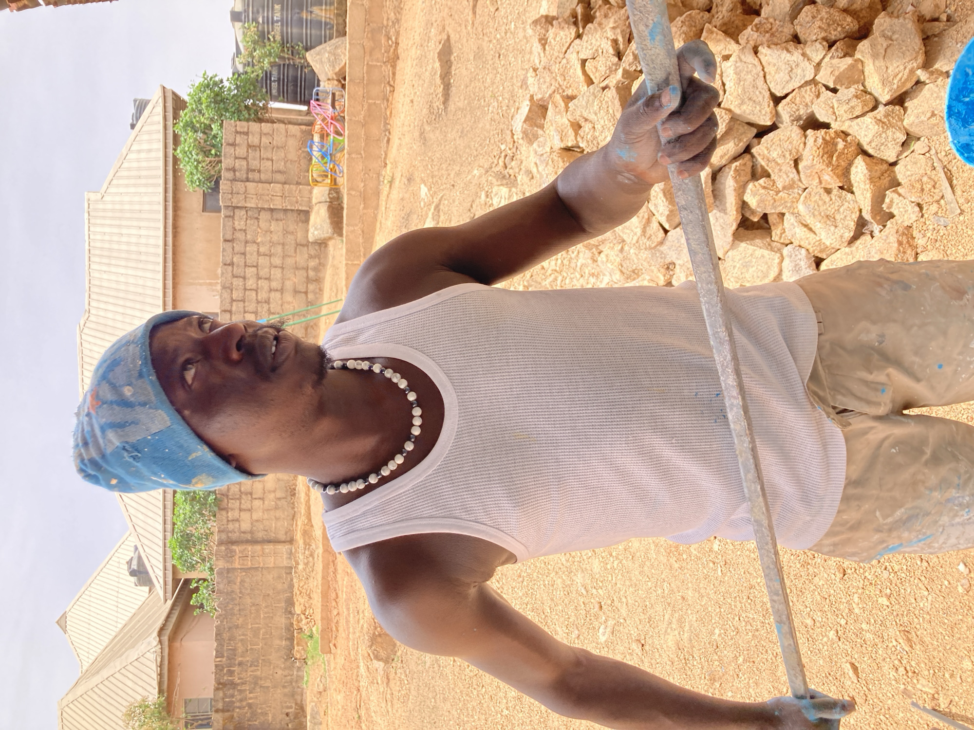 A brightly lit, outdoor, full-body shot captures a man engaged in painting or paint preparation in what appears to be a work area in Jos, Nigeria. The man, with dark skin, wears a white ribbed tank top, a light blue paint-splattered head covering, and light-colored, paint-stained trousers. A white and dark beaded necklace is visible around his neck. Blue paint splatters his hands, arms, and clothing, indicating his current activity. He is holding a long, blue paint-covered stirring rod with both hands, looking upwards and slightly to his left with a neutral, focused expression.
In the foreground, a blue container is partially visible near his right foot. Behind him, a large pile of light-colored, rough-hewn rocks forms a backdrop. The ground is dry, dusty, and sandy. The background includes buildings with light-colored metal roofs and brick/block walls. A prominent black water tank, labeled "ANGEL TANK", is mounted on a stand above one of the structures. Green foliage and colorful plastic rings are also visible in the distance. The bright, clear lighting suggests a sunny daytime setting. The scene portrays a practical, utilitarian environment.