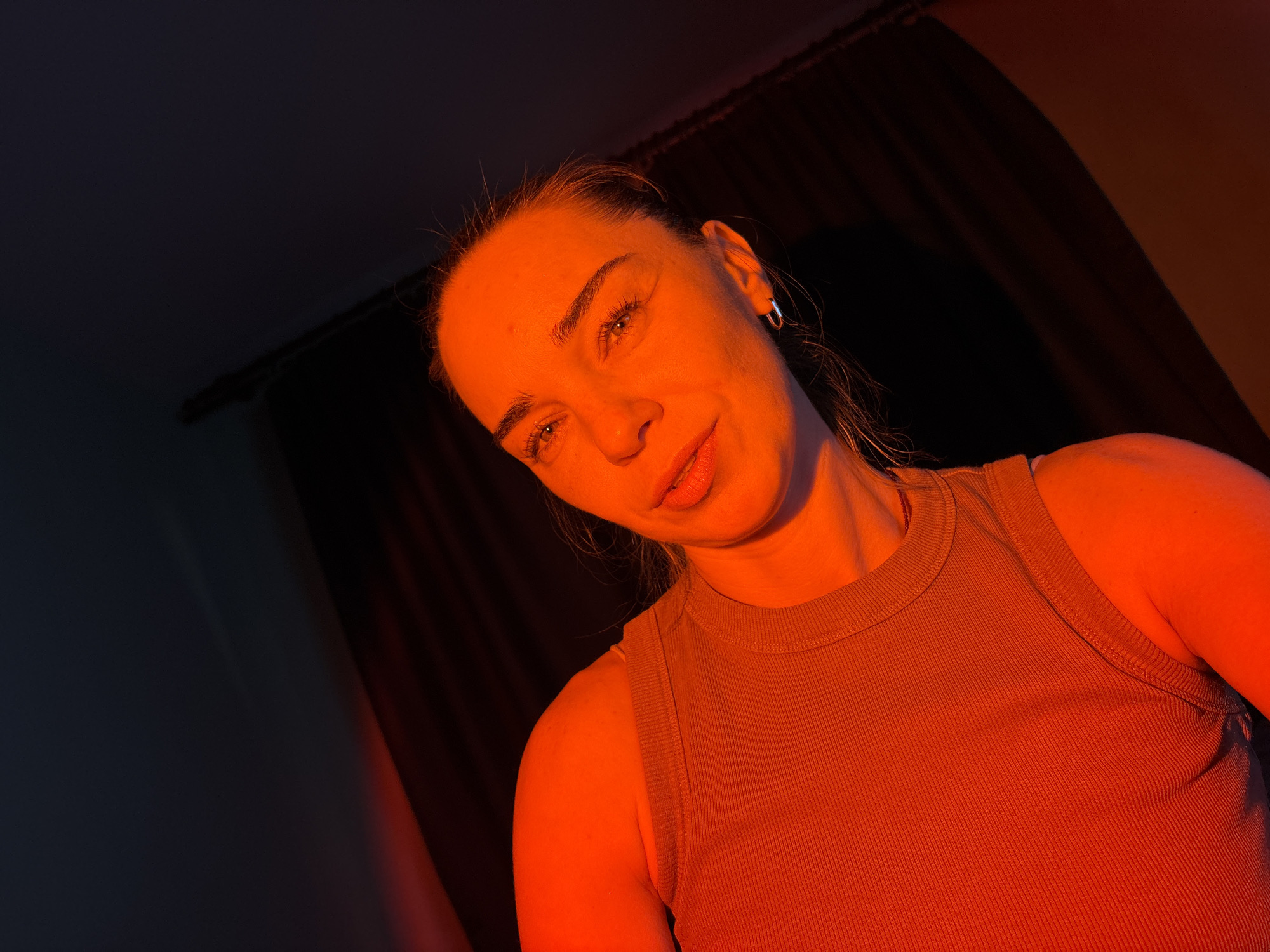 A close-up, low-angle shot shows a woman illuminated by red lighting against a dark background. The woman is looking up and to her right, with her head tilted. She has blonde hair pulled back, and is wearing a tan tank top and a small silver hoop earring in her left ear. Her makeup includes red lipstick and mascara. The background appears to be a dark room with a curtain. The lighting casts strong shadows and highlights on her face and body, giving the image a dramatic feel.