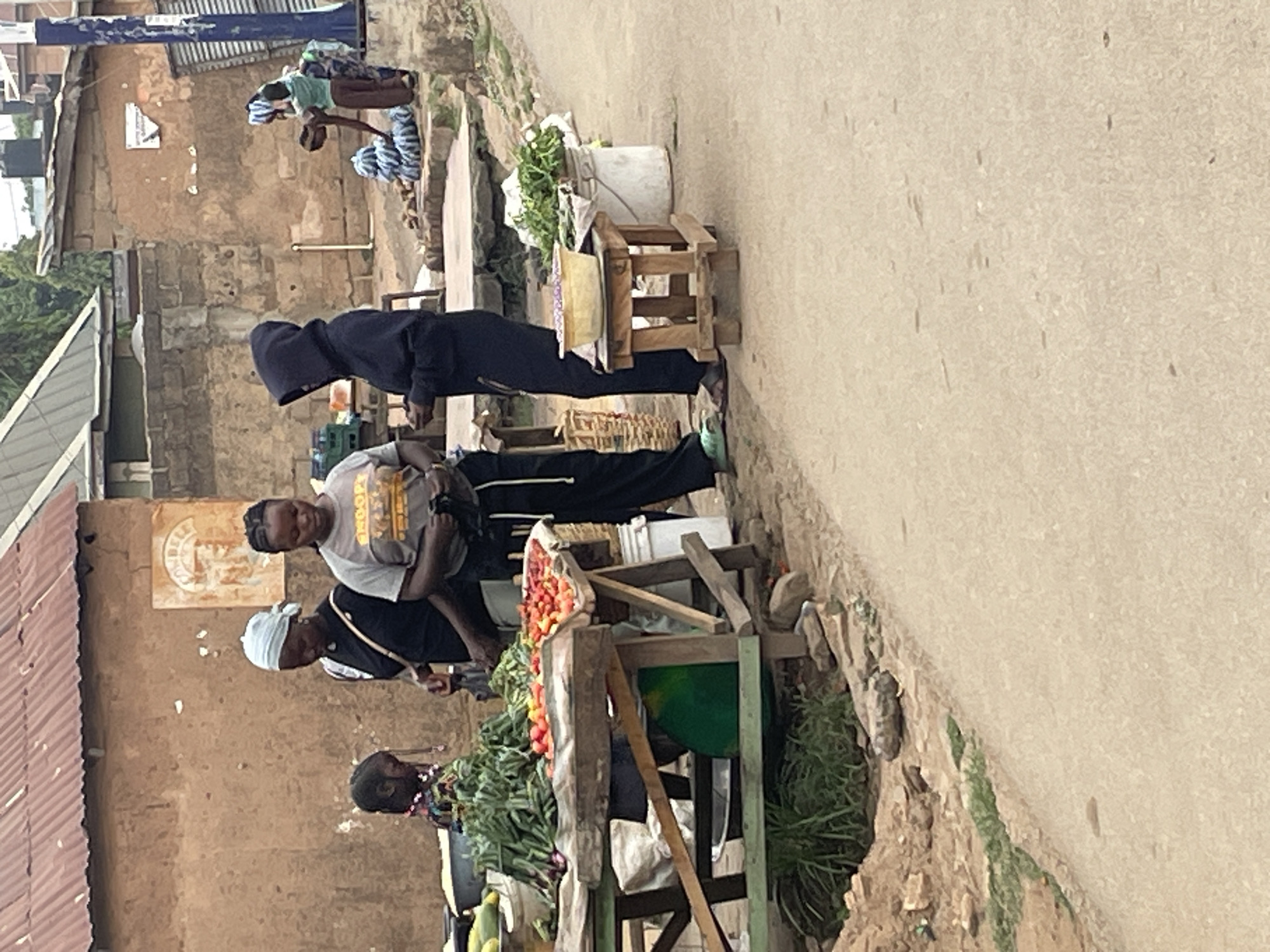 The image depicts an outdoor scene in Jos, Nigeria, showcasing a roadside vegetable stand. Three individuals, including two women and one man, are observed near the stand. The women are positioned close to the produce, seemingly engaged in a transaction or assisting at the stand. The man, further away, appears to be observing the scene. The stand exhibits a variety of fresh produce, including tomatoes, greens, and other vegetables. In the background, other people are observed in a more distant area of the scene. The environment is characterized by buildings with visible wear and tear, situated along a road or pathway. Overall, the image captures a glimpse of everyday life and commerce in Jos, Nigeria.