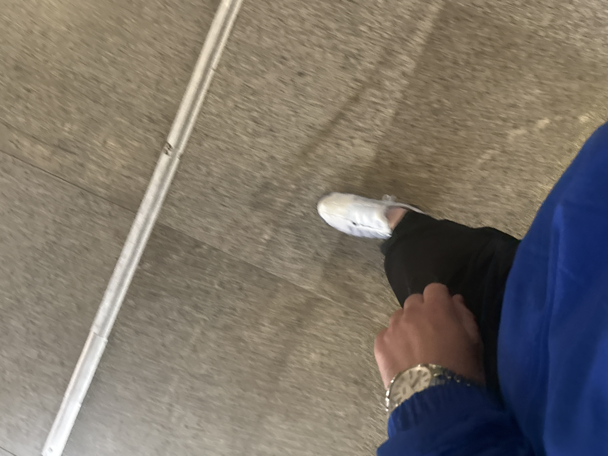 The image is a first-person, downward-looking shot, capturing a person's lower body and left arm as they stand or move indoors. The person wears a vibrant royal blue long-sleeved top or jacket, black pants, and white athletic shoes. A silver-toned watch with a metal link bracelet is visible on their left wrist. The flooring is a light grey-brown with a subtle horizontal grain or texture, suggesting laminate or a similar surface. A long, light grey or silver-colored strip, possibly a rail or guide, runs diagonally across the floor from the upper left to the lower right. Small, indistinct dark markings, which could be numbers or letters (resembling a '2'), are barely discernible on this strip but are illegible due to blur and low resolution. The watch face also has text, but it is unreadable. The image exhibits a slight motion blur, indicating either the subject or the camera was in motion. The setting is an indoor public space, potentially in Sofia, Bulgaria.