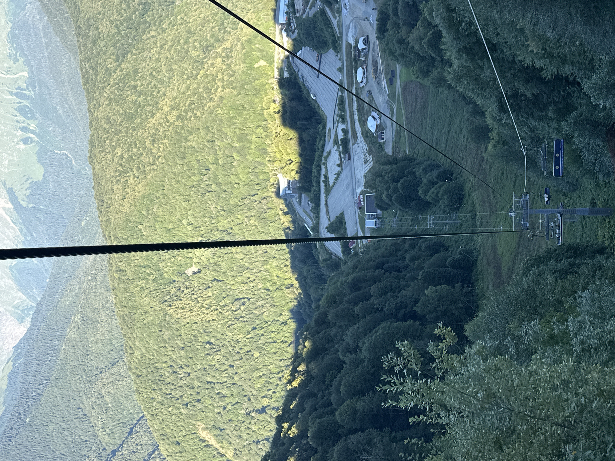 The image shows a cable car descending a mountainous slope covered in dense green trees, with a small settlement visible in the distance. The cable car, with multiple empty seats, hangs from a thick black cable that stretches diagonally across the frame. Sunlight casts a strong shadow across the lush forest, highlighting the contours of the mountains. In the lower portion of the image, the settlement appears to consist of several buildings and large, empty parking lots, suggesting a resort or infrastructure area within the natural landscape. The background reveals layered mountain ranges, some of which are partially obscured by haze. The overall scene depicts a scenic view of a forested mountain resort area, likely in Esto-Sadok, Russia, during daylight.