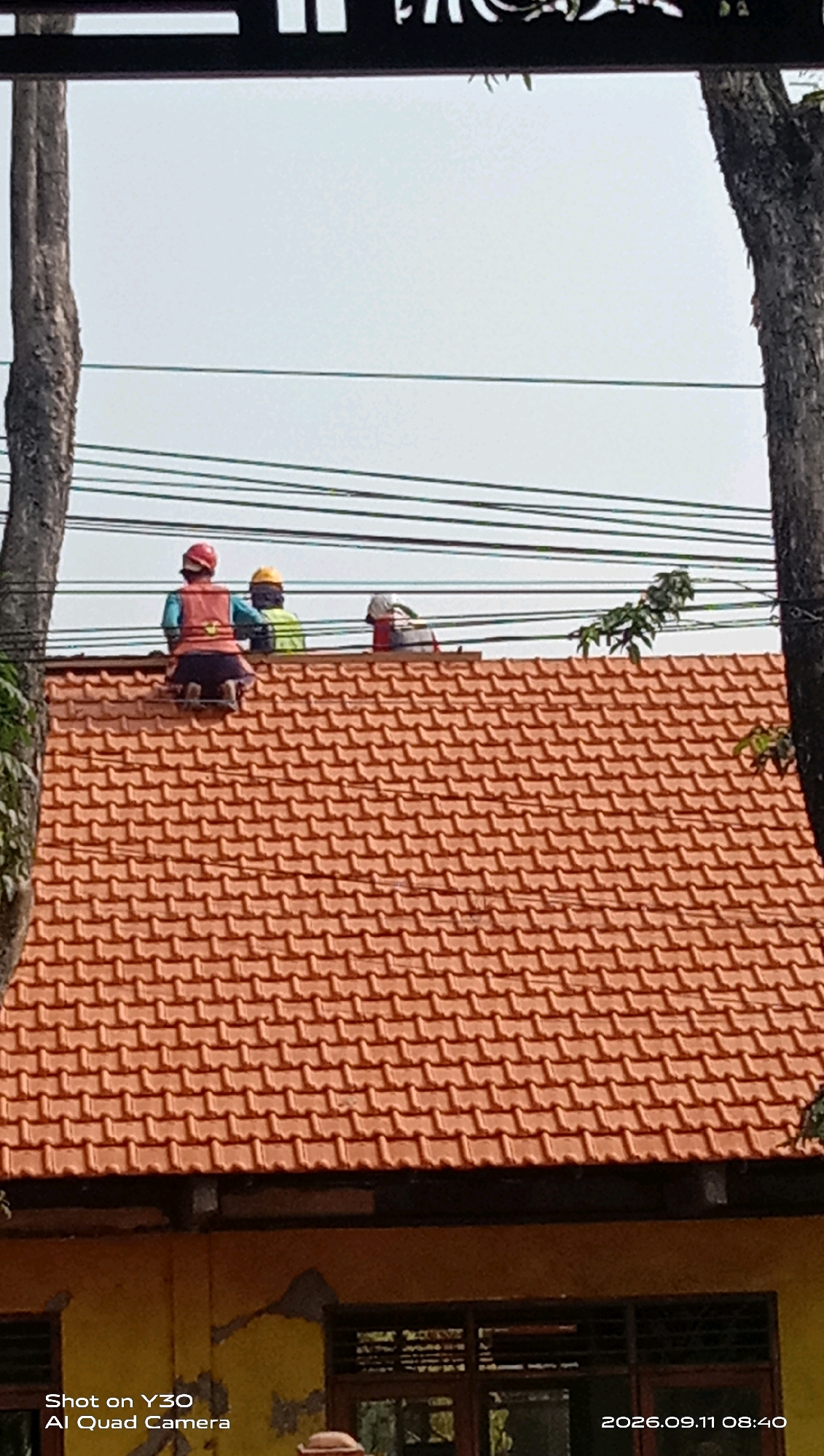 Three individuals wearing hard hats are situated on an orange-tiled roof. The person on the far left is kneeling, wearing a red vest and dark pants. To their right, another individual is standing, wearing a green vest, and further to the right, a third individual is also standing, wearing a red and white patterned shirt. They appear to be working near power lines that stretch across the frame above the roof. The background consists of a light-colored sky and two large trees framing the scene on the left and right. The building's facade below the roof is yellow with visible signs of wear and tear, and windows with dark frames are present. The timestamp "2026.09.11 08:40" is visible in the lower right corner, alongside the text "Shot on Y30 AI Quad Camera." The location is specified as Munggugianti, Indonesia.