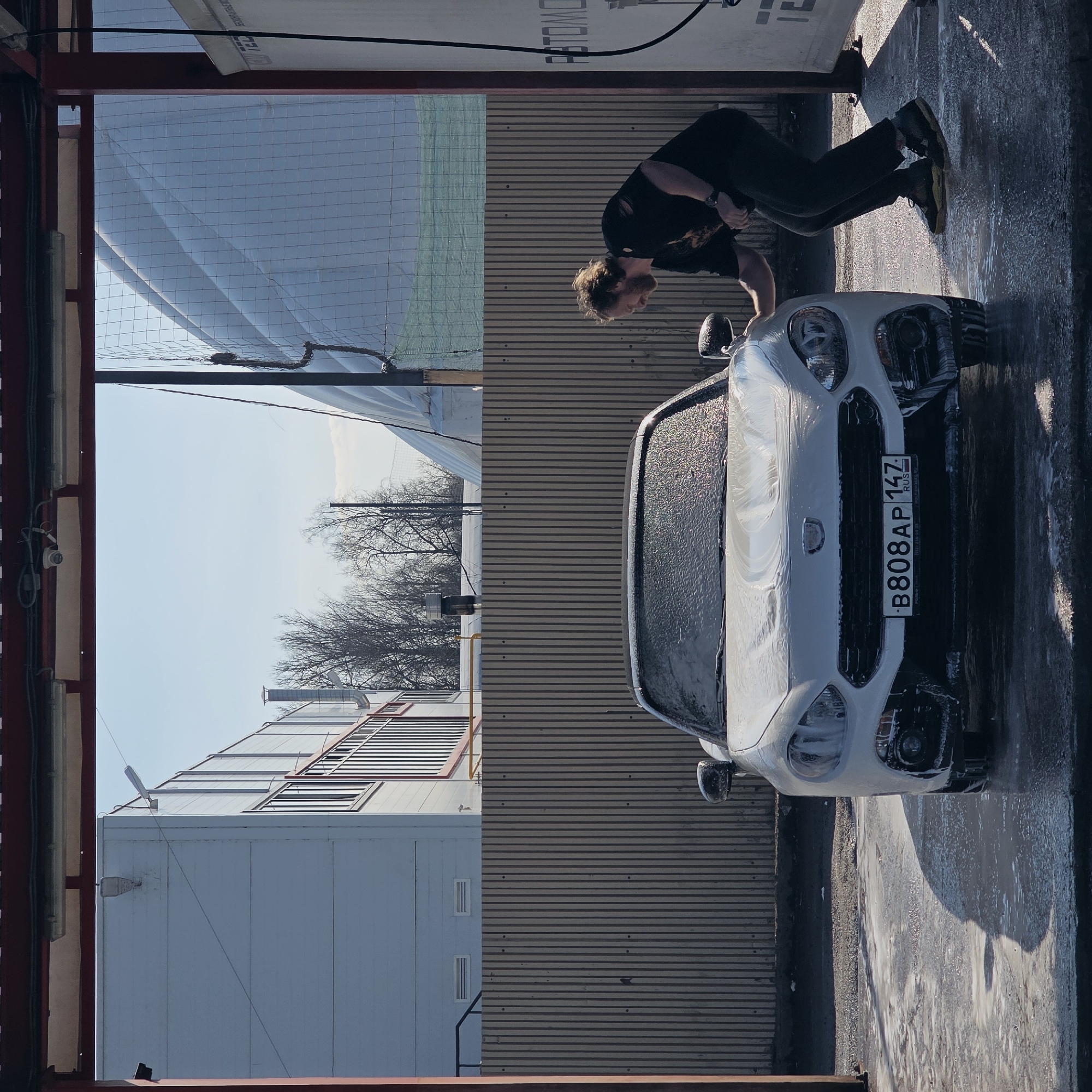 A medium shot depicts a man cleaning a white car at a car wash, presumably in Saint Petersburg, Russia, given the license plate number "B808AP 147" on the car's front. The man, dressed in dark clothing and tan work boots, is bent over the hood of the car, his hands possibly holding a sponge or cloth. The car is covered in soap suds and water, indicating that it has recently been washed. The car wash appears to be indoors, with corrugated metal walls, a ceiling with a wire mesh, and an industrial building visible in the background.