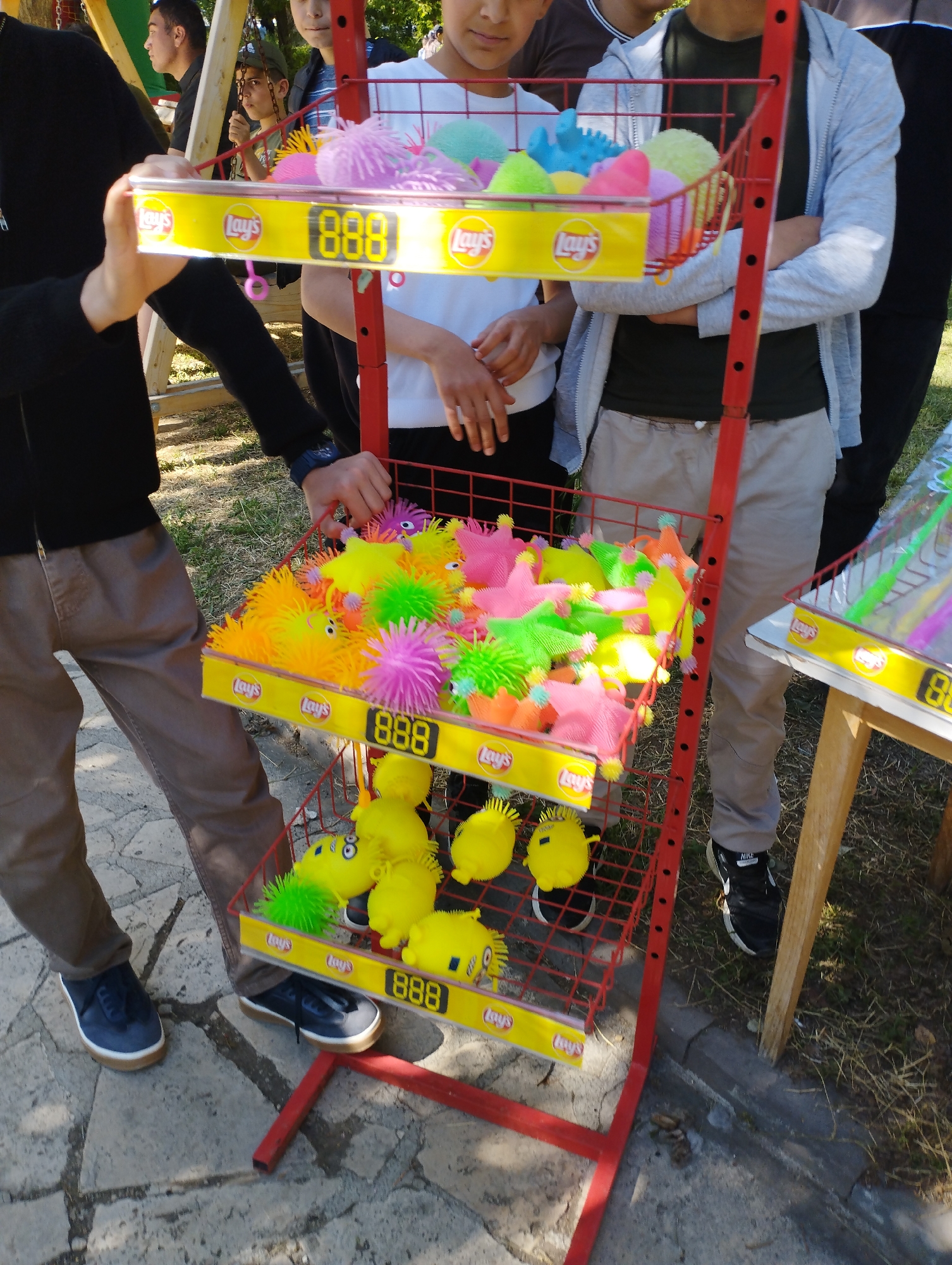 In an outdoor setting, possibly in Yeghegnadzor, Armenia, a red metal display stand with three shelves filled with colorful, spiky toys resembling sea creatures is the focal point. The stand has "Lay's" branding and the number "888" on each shelf's front panel. A group of children, primarily boys, are gathered around the display. One boy in a black jacket and brown pants is reaching toward the top shelf. Another boy in a white shirt stands in front of the display. A third boy in a gray jacket and khaki pants is to the right, with his arms crossed. The ground appears to be a mix of stone paving and grassy areas. A small wooden table with more toys is visible to the right.