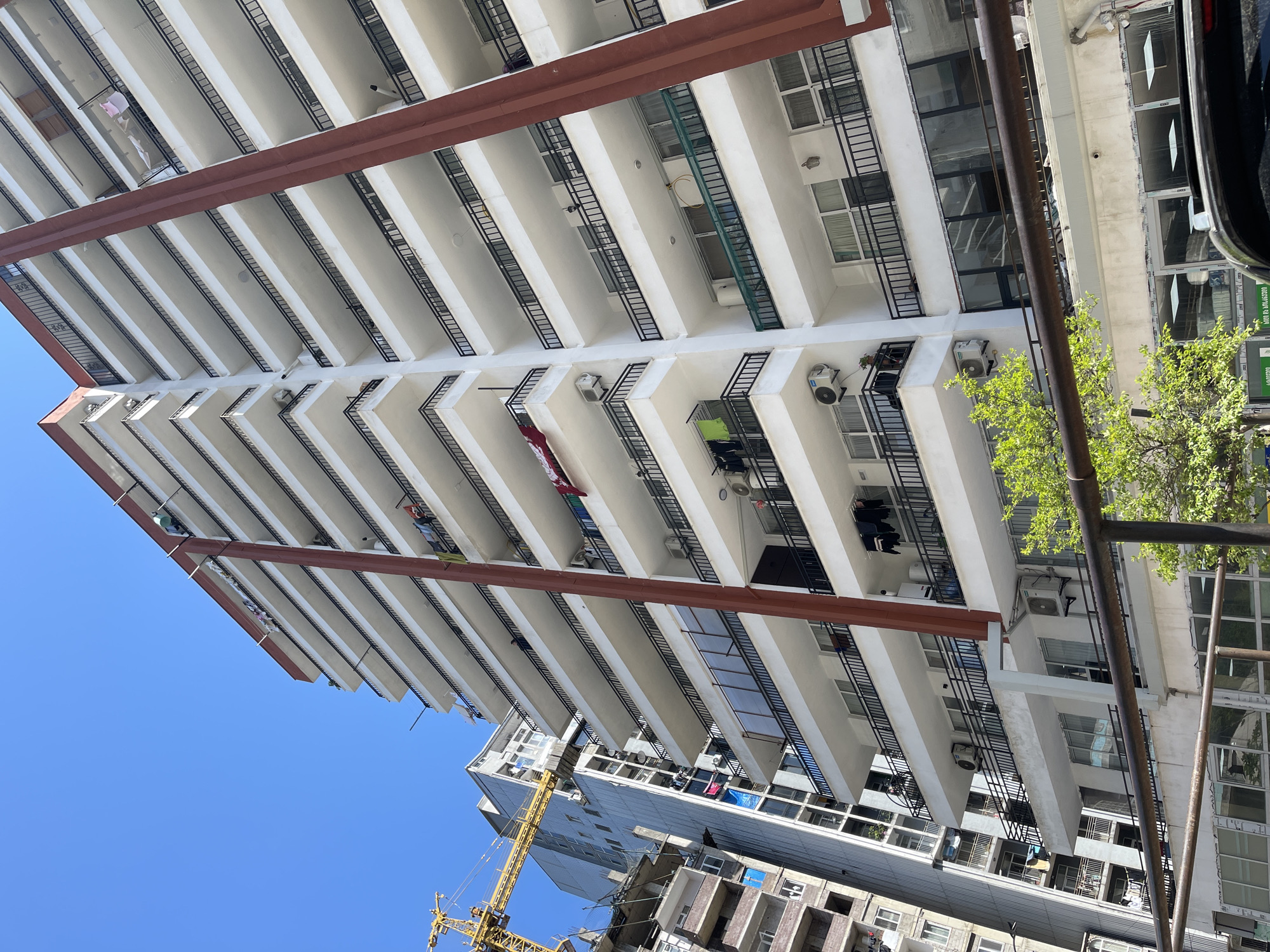 Looking up at a tall, modern apartment building in Batumi, Georgia. The building has numerous balconies, some with air conditioning units and hanging laundry. A construction crane is visible in the background, indicating ongoing development in the area. The sky is clear and blue.
