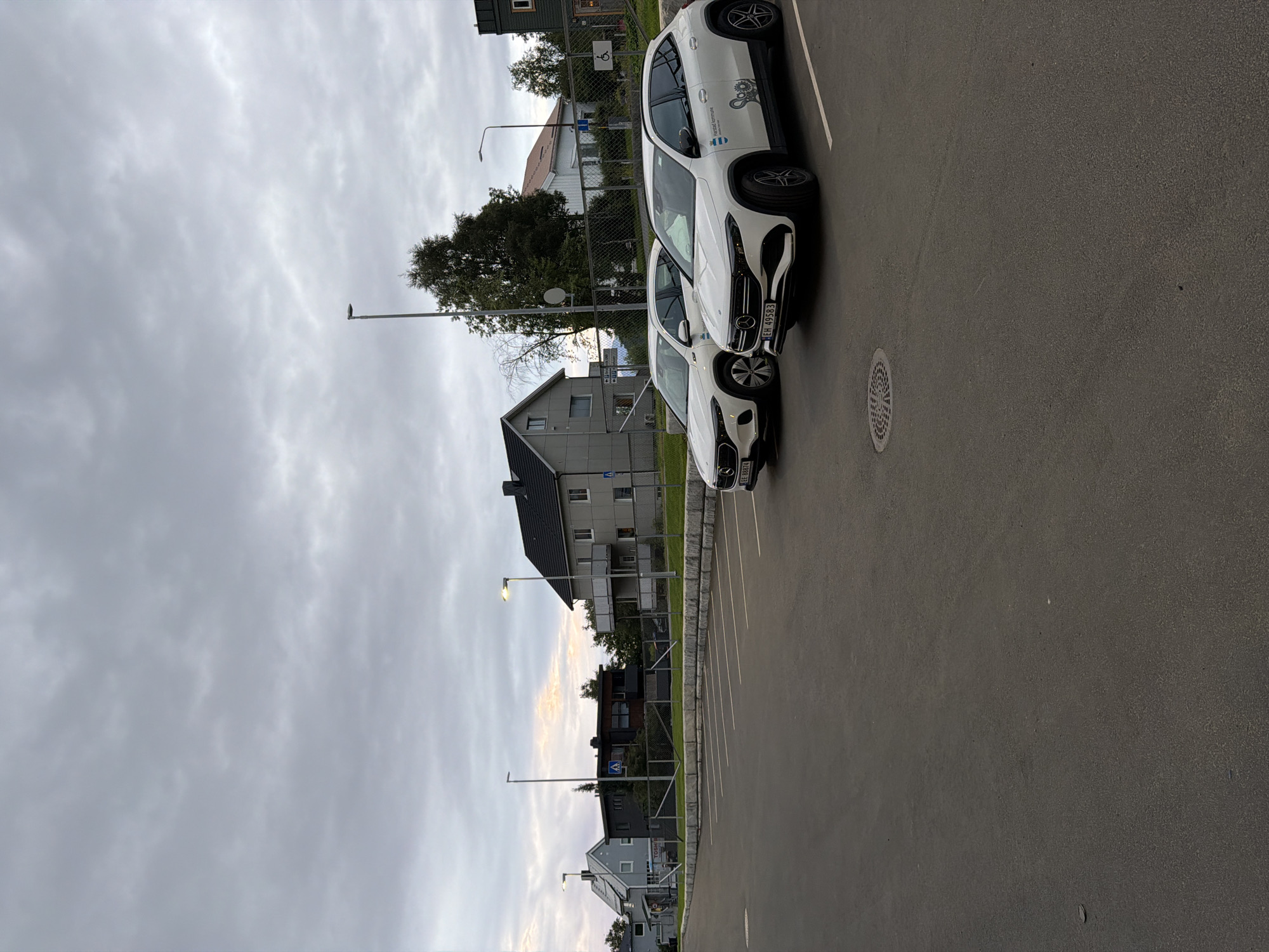 The image depicts an outdoor parking lot in Harstad, Norway, under a cloudy sky. Two white Mercedes-Benz EQA electric vehicles are parked side-by-side in the foreground. The vehicle on the right displays "Harstad Kommune" and a logo with gears on its door, indicating it belongs to the municipality. The vehicle on the left has license plate EE 88874, and the vehicle on the right has license plate EH 49583. Behind the cars, a chain-link fence separates the parking lot from a sidewalk and a row of buildings. These buildings appear to be residential or commercial, with a mix of modern and traditional architecture. Streetlights are visible along the sidewalk, and trees are scattered throughout the scene. The ground of the parking lot is asphalt, marked with white parking lines and a circular manhole cover. The overall atmosphere is overcast and subdued, suggesting it might be late afternoon or early evening.