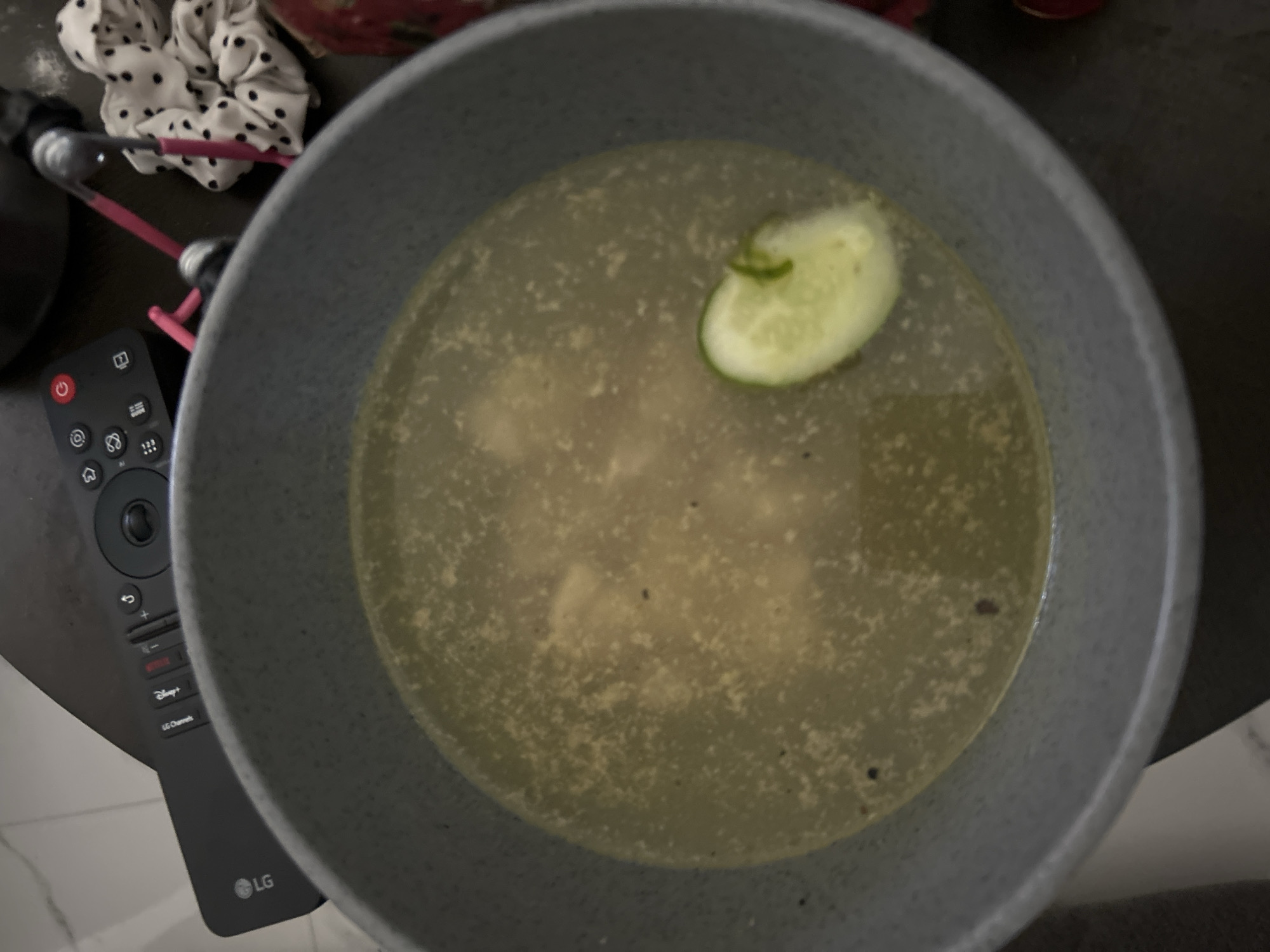 A close-up overhead shot shows a light gray bowl filled with a yellowish broth. A single slice of cucumber floats in the broth, with some small dark specks also visible throughout the liquid. The background is blurred, but appears to be a dark surface, possibly a table, with a black LG remote control and other out-of-focus objects. The scene is not located in Sukawati, Indonesia.