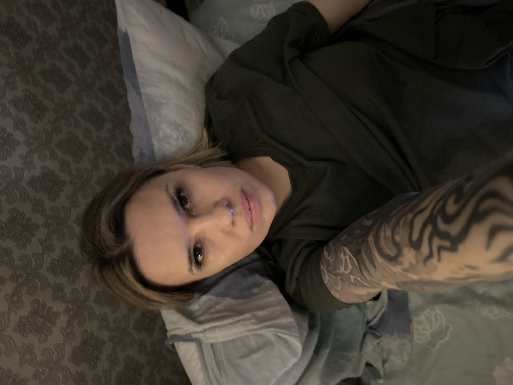 An indoor scene depicts a single female individual lying on a bed, captured in a self-portrait. The subject, an adult female with blonde/light brown hair, stares directly at the camera. She has visible eyelashes and a silver-colored septum piercing. She is wearing a dark olive green short-sleeved t-shirt. Her left arm, extended towards the camera, is covered by a full sleeve tattoo featuring black and grey abstract or tribal patterns. She rests on light-colored pillows, one with a subtle floral or leaf pattern, and light blue and white patterned bedding is visible. The background consists of a wall covered in dark grey/brown wallpaper with a repeating damask or floral motif. The specific location in Rostov-on-Don, Russia, cannot be determined from the visual evidence within the image.