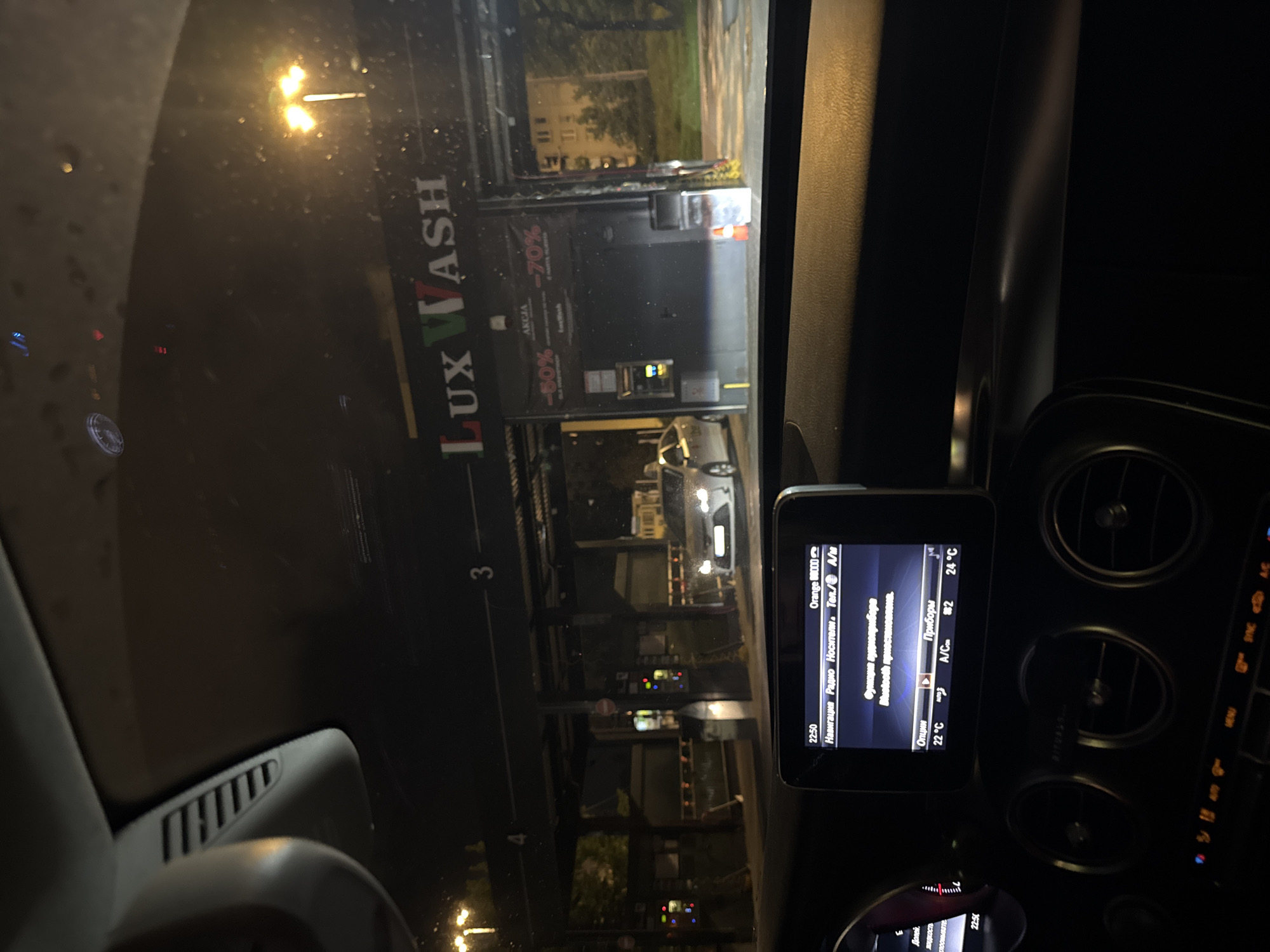 The image is taken from inside a car at night, looking out at a car wash station. The car wash has a sign that says "LUX WASH" in large letters, with "AKCJA" (Action/Sale in Polish) and promotional prices like "-50%" and "-70%" displayed. Several cars are visible, some entering or exiting the car wash bays. The interior of the car shows a dashboard with a screen displaying information, including time "22:50", navigation and radio options, and climate control settings. The environment appears to be urban, with streetlights and buildings visible in the background. The overall scene suggests the car is waiting in line to enter the car wash.