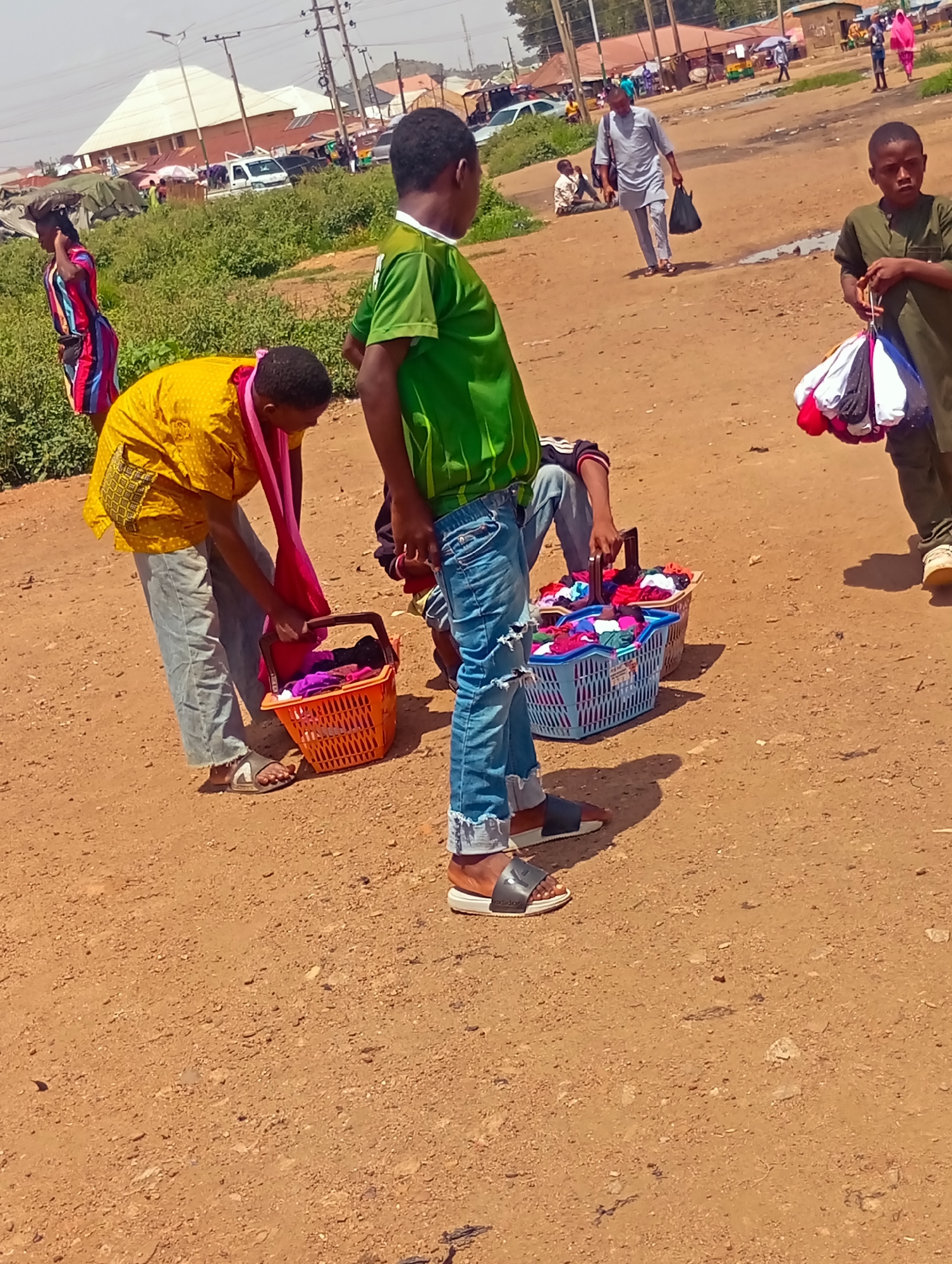 In Bukuru, Nigeria, a street scene unfolds under a bright sky with a dirt road and scattered vegetation. In the foreground, a young boy in a green shirt and ripped blue jeans stands holding a bundle of colorful items, possibly socks or small garments, for sale. To his left, a woman in a yellow top and pink scarf sorts through an orange basket filled with clothing. Further back, another person sits on the ground next to a blue basket overflowing with similar colorful items. To the left, a woman in a vibrant, striped dress walks away from the camera, carrying something on her head. In the background, various structures and vehicles suggest a bustling marketplace environment, with more people and stalls visible. The overall scene depicts a moment of commerce and daily life on a dusty Nigerian street.