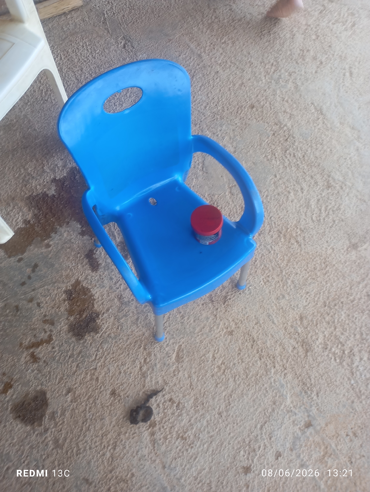 A bright blue plastic child's chair is positioned on a rough, concrete ground surface, possibly outdoors in a yard or patio. The chair features a circular cutout in its backrest and silver metal legs with blue plastic caps on the feet. A small, red-lidded cylindrical container rests on the seat of the chair. The ground is uneven and shows some dark stains, suggesting it might be damp or dirty. In the upper left corner of the image, a portion of a white plastic chair is visible. The scene is illuminated by natural light. The image also contains a timestamp and text overlay indicating "REDMI 13C" and "08/06/2026 13:21". While the image itself does not provide direct evidence of the city of Jos, Nigeria, the presence of typical building materials and the general environment could be consistent with an urban or semi-urban setting in that region.