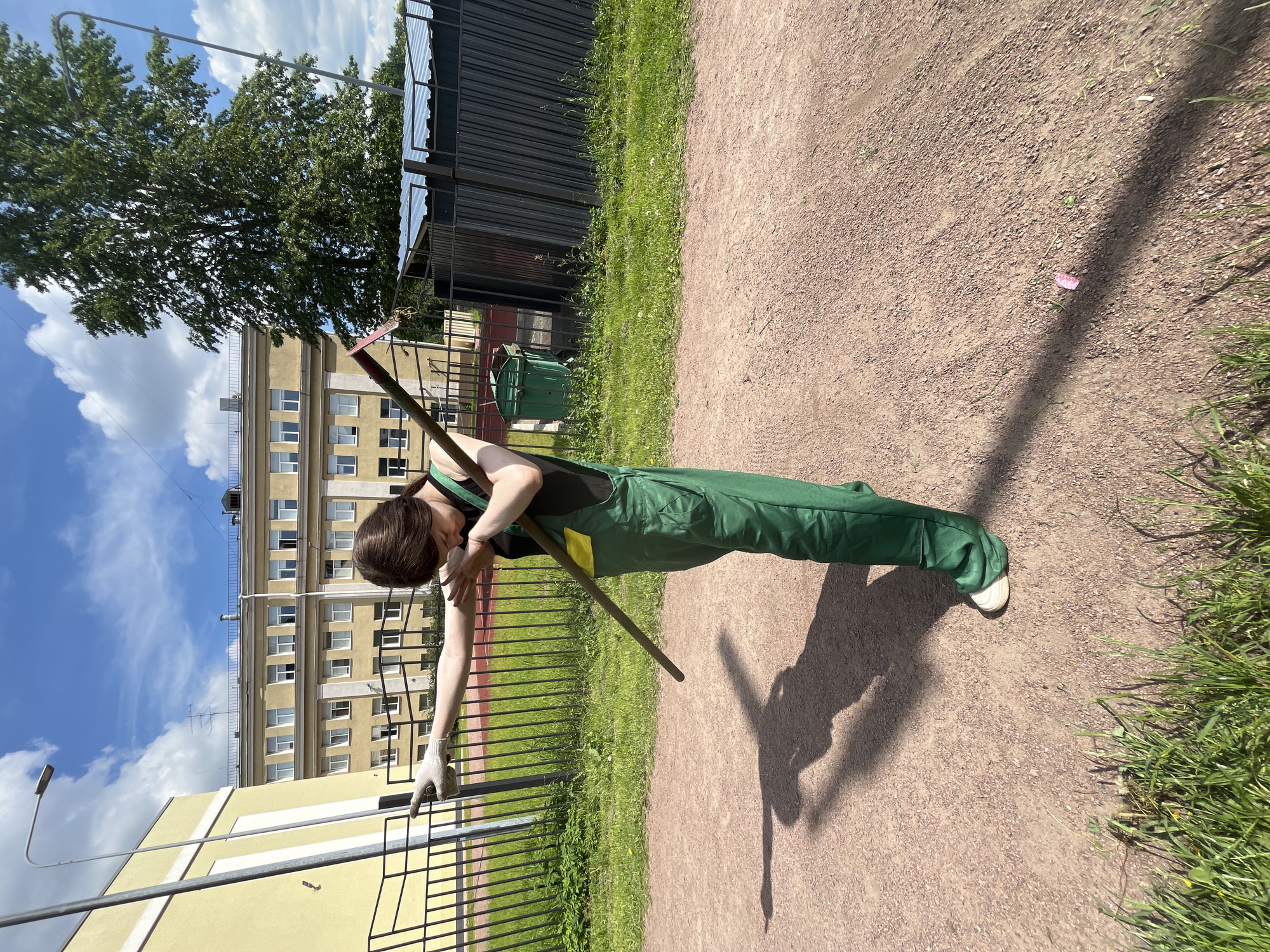A young woman wearing green overalls and white gloves stands outdoors, holding a long pole with a red tip. She is performing the "dab" pose, with one arm extended horizontally and the other bent across her chest. She is looking down, with her head bowed. The background features a yellow building with multiple windows, a black metal fence, a green trash bin, and a large tree. The ground is covered with reddish gravel. The sky is blue with scattered white clouds. This scene appears to be in an urban environment, possibly a schoolyard or park in Saint Petersburg, Russia, given the architectural style of the building.