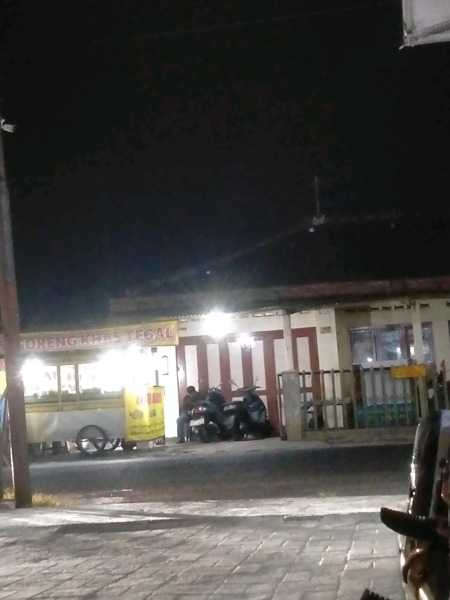 This is a nighttime exterior shot of a street scene in the city of Kasihan, Indonesia. The foreground shows a brick-paved sidewalk with dappled lighting. The midground features a street with a food cart on the left side, displaying a banner that reads "GORENG KHAS TEGAL" and "MIE GORENG AYAM". A person is seated on a motorcycle to the right of the food cart, with another motorcycle parked behind them. A low wooden fence with vertical posts separates the street from the building in the background. The building has a light-colored facade with red and white striped vertical panels and windows. The sky above is dark, indicating it is nighttime.