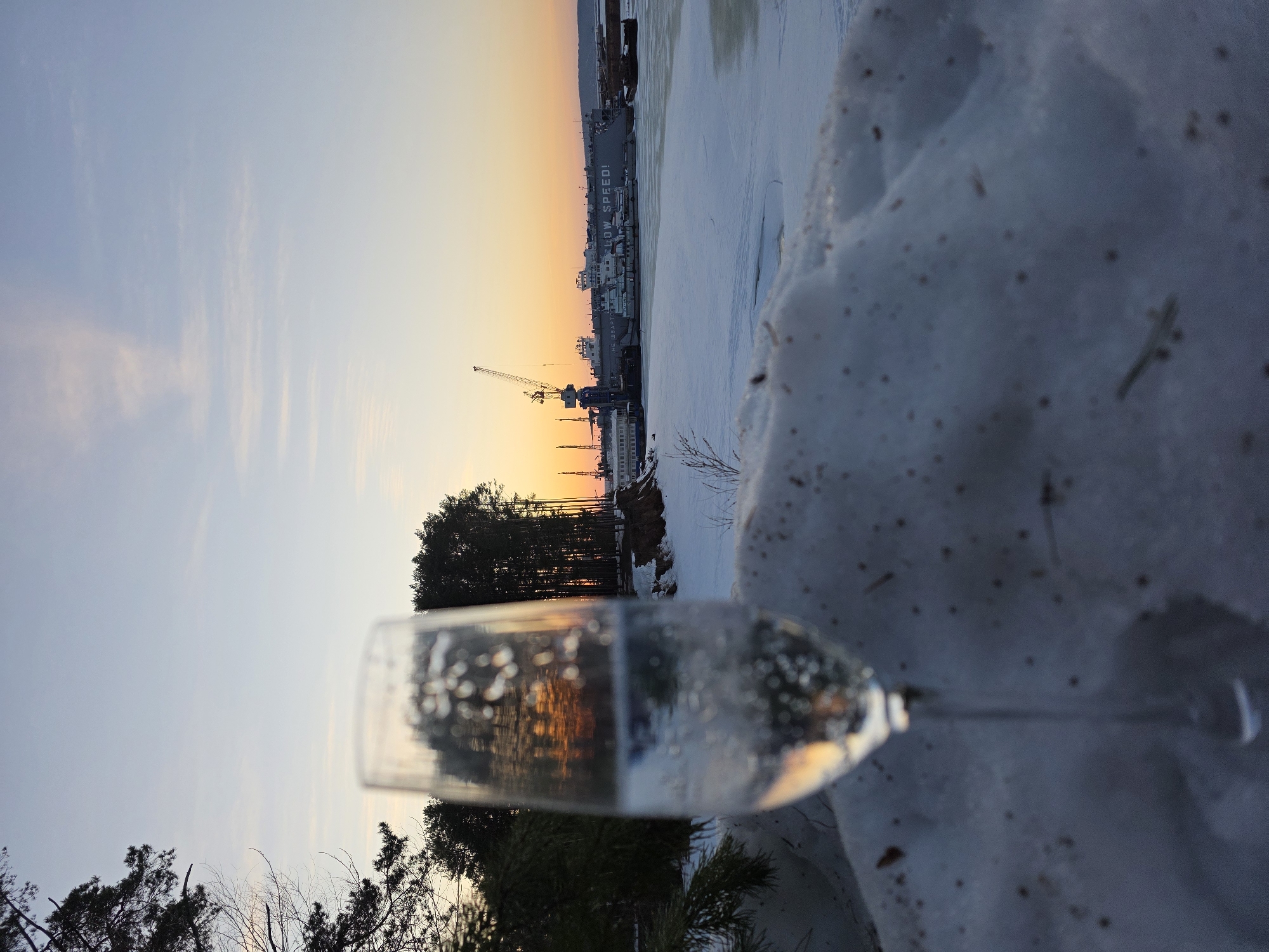 The image shows a winter scene at sunset in Chaikovsky, Russia. In the foreground, a champagne flute partially filled with liquid is placed on a snowy surface. The liquid reflects the colors of the sunset and nearby trees. In the background, a frozen river leads to a cityscape with buildings and cranes, silhouetted against the orange and yellow sky. The "SLOW SPEED!" ship is visible in the river. Bare trees are also present in the foreground, framing the view.