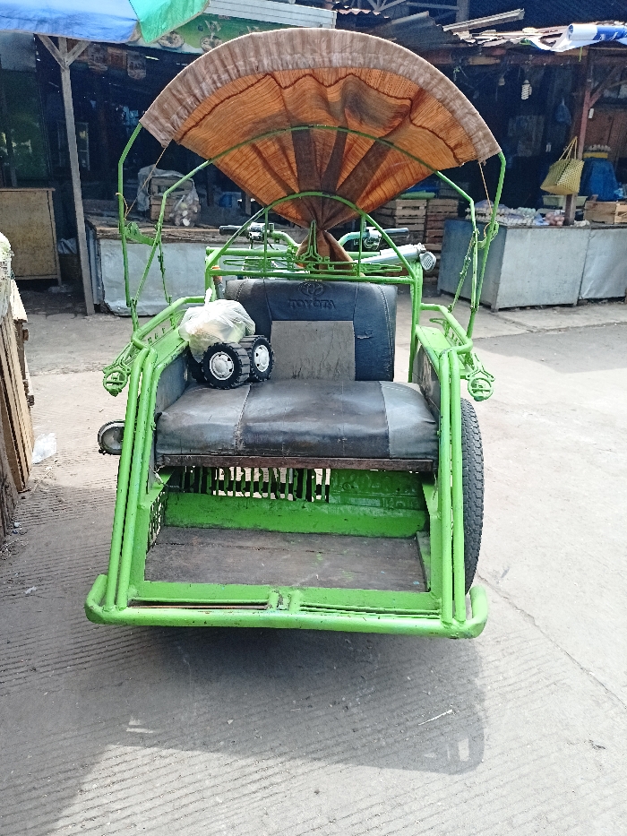This image features a bright green pedicab, also known as a cycle rickshaw or trishaw, parked outdoors. The pedicab's frame is made of sturdy green metal tubing. It has two passenger seats in the front, upholstered in black and grey, with the word "TOYOTA" visibly embroidered on the backrest of the rear passenger seat. A small toy vehicle, possibly a toy tank or toy with tracks, and a white plastic bag are resting on the passenger seats. Above the seats, there is a fan-shaped canopy made of woven, light brown material, providing shade. A single wheel is partially visible on the right side of the pedicab.
The setting appears to be a market area or a street lined with small stalls. In the background, there are various structures made of wood and what look like corrugated metal or simple coverings, some draped with blue tarpaulins. Other goods, possibly produce or market items, are stacked or displayed on these stalls. The ground is a concrete or paved surface, showing distinct shadows, indicating bright daylight, likely midday or early afternoon, under sunny weather conditions. No people are visible, and no specific activity is taking place; the pedicab appears to be stationary, possibly awaiting passengers or simply parked. The style of the vehicle and surroundings is consistent with the location context of Mojosari, Indonesia.