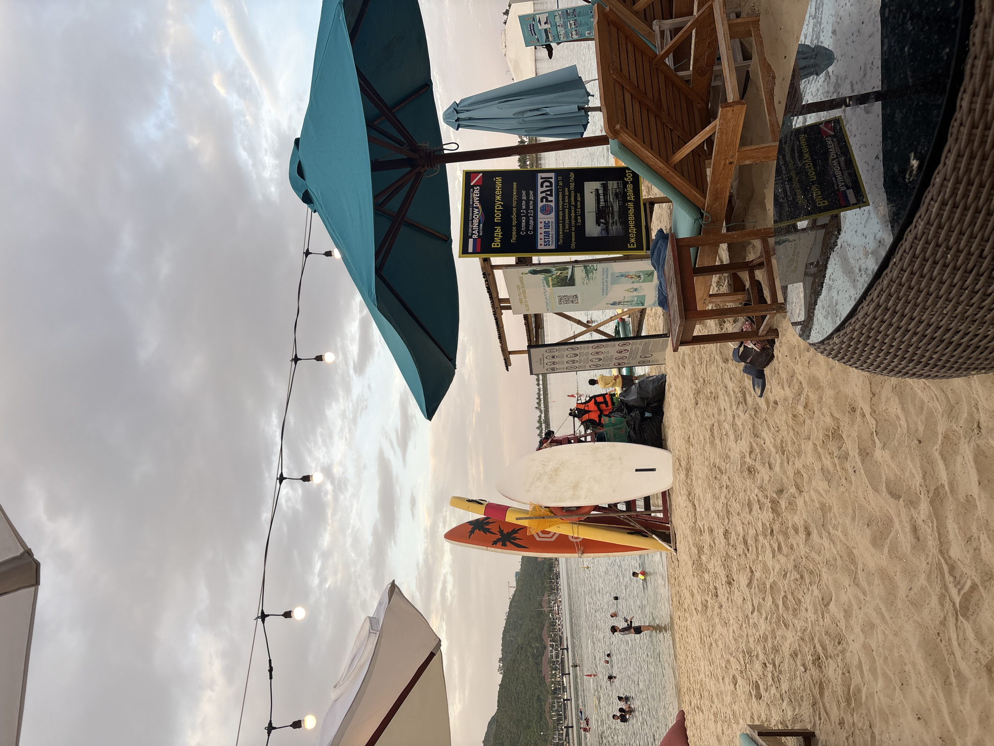 The image shows a beach scene on a cloudy day, likely in Nha Trang, Vietnam, given the presence of surfboards and the general ambiance. In the foreground, a woven coffee table with a reflective surface sits on the sand. Behind it, a wooden lounge chair is positioned, partially under a large teal umbrella. To the left, a small wooden side table is placed next to the lounge chair.
Further back, several surfboards are propped up against a red frame. Next to them, a man in a bright orange life vest stands next to several black garbage bags, possibly working. A blue upright umbrella stands further to the right.
The middle ground features a body of water with several people swimming and wading. In the background, a lush green hill rises, dotted with buildings and some construction. The sky is overcast with diffused light. String lights with glowing bulbs are strung across the upper portion of the image, adding a festive touch. Signs advertising diving services, including "RAINBOW DIVERS" and "PADI," are visible, indicating recreational activities are offered.
