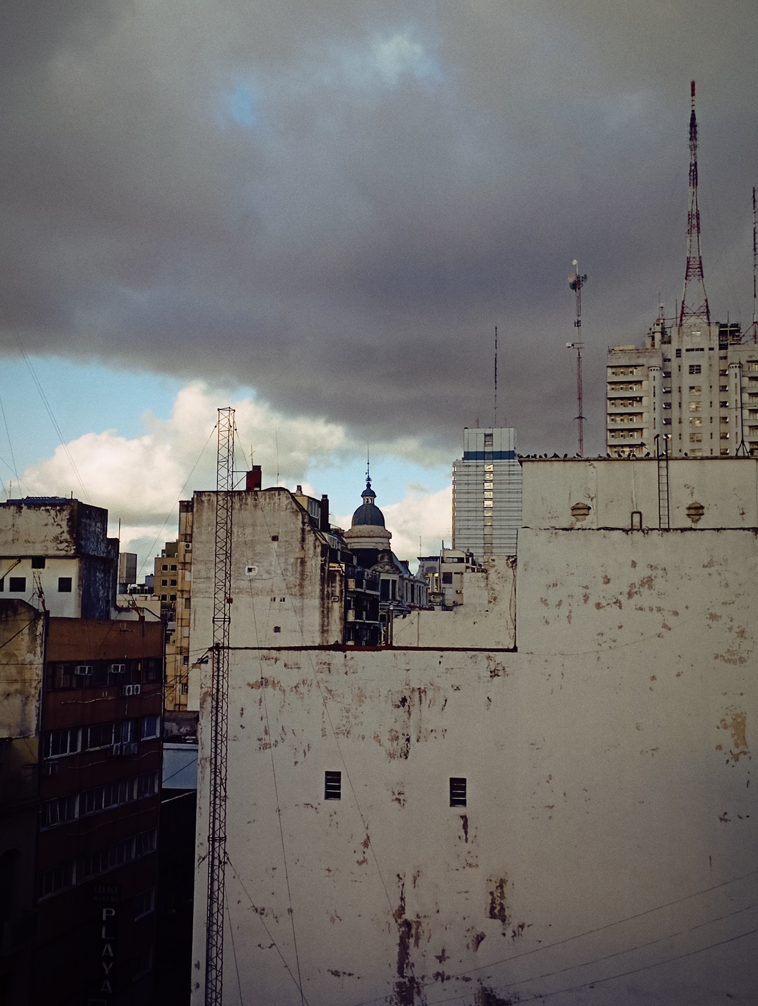 The image shows a cityscape under a cloudy sky, likely taken in Buenos Aires, Argentina, judging by the architectural style and the presence of structures typical of the city. Several buildings of varying heights are visible, constructed with a mix of materials. The buildings in the foreground have weathered, light-colored facades. A communication tower is prominent on top of one of the higher structures. Dark, ominous clouds dominate the upper portion of the frame. The angle suggests the photo was taken from an elevated position.