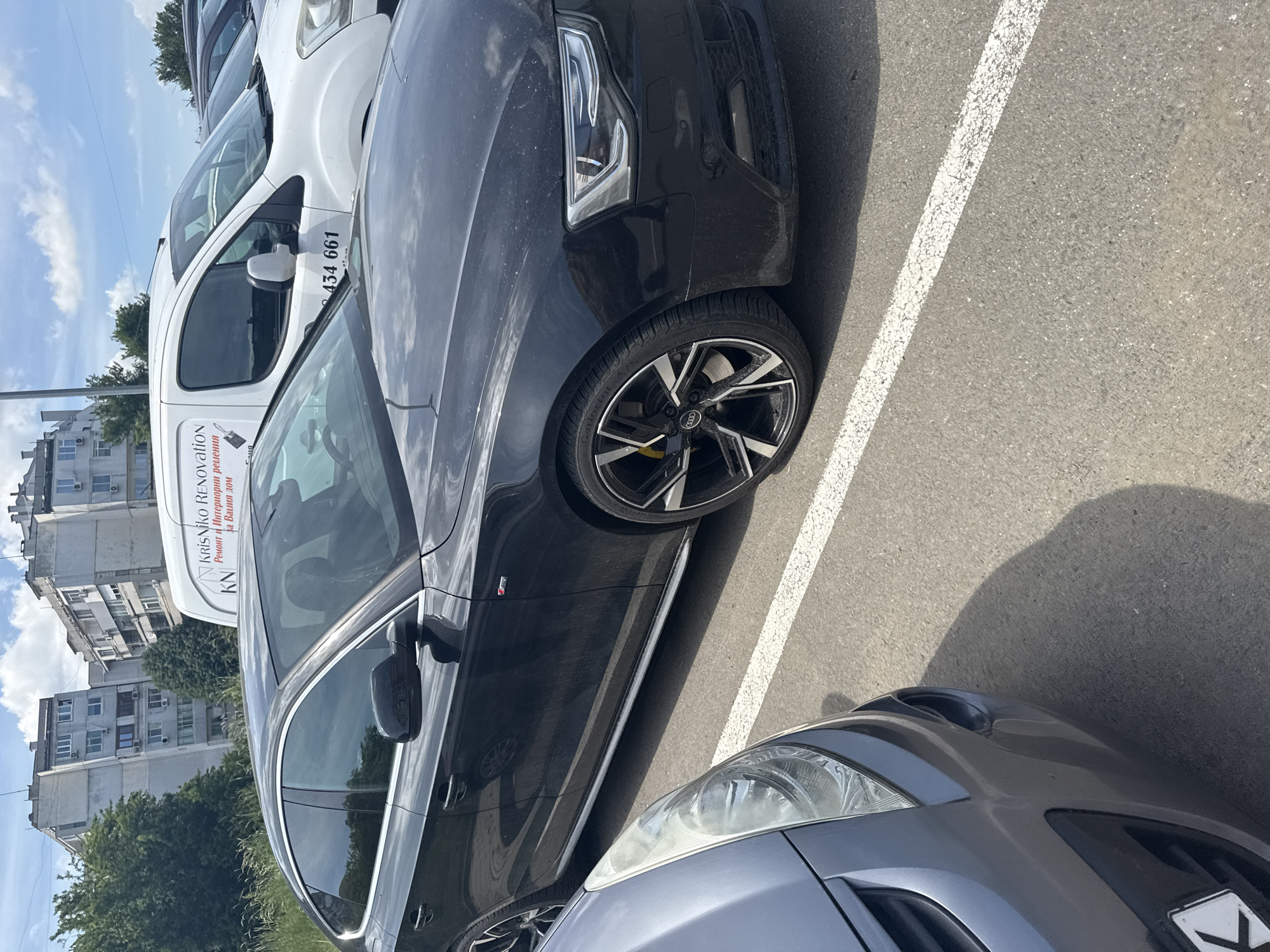 The image shows a street scene in Burgas, Bulgaria. A black Audi car is parked in a parking space defined by white lines on an asphalt surface. To the left of the Audi is a white van with "KRISIKO RENOVATION" and a Bulgarian phone number "434 661" printed on its side. The van also has text that says "Ремонт и Интериорни решения за Вашия дом." To the right of the Audi is a gray car, with the front right corner visible. The background shows a residential area with apartment buildings and trees under a partly cloudy sky. The photo appears to have been taken during the day.