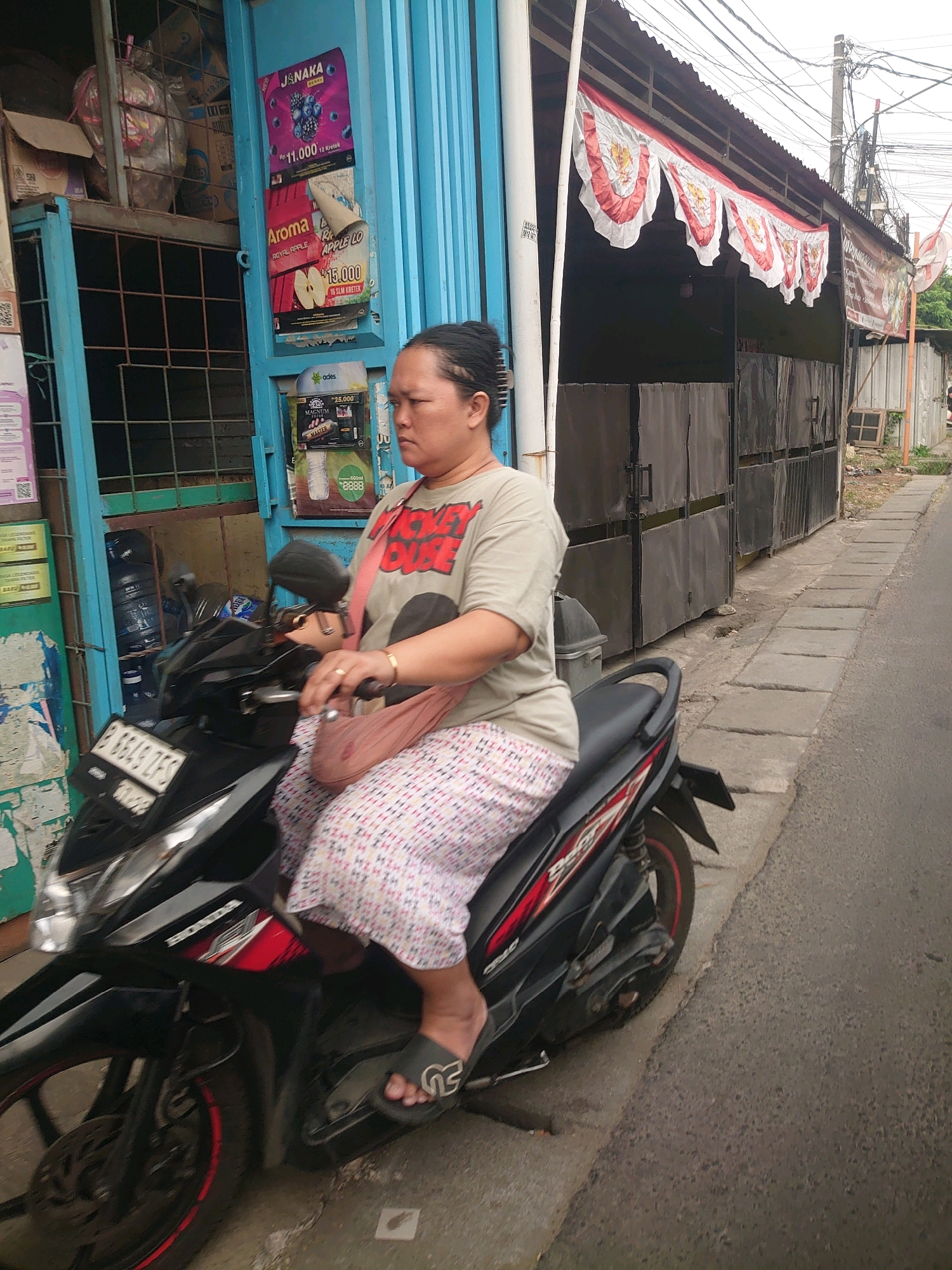 A woman with dark hair pulled back is sitting on a black motorcycle, facing right. She is wearing a grey t-shirt with a red graphic of Mickey Mouse and a white and pink patterned skirt. She has a pink bag slung over her shoulder. The motorcycle has a license plate that reads "B 6849 ZFS". The woman is on a street in Depok, Indonesia, with a building with blue shutters and metal doors to her left. Red and white banners with bird emblems are hanging from the awning of the building. The street has a concrete sidewalk and is lined with power lines overhead.
