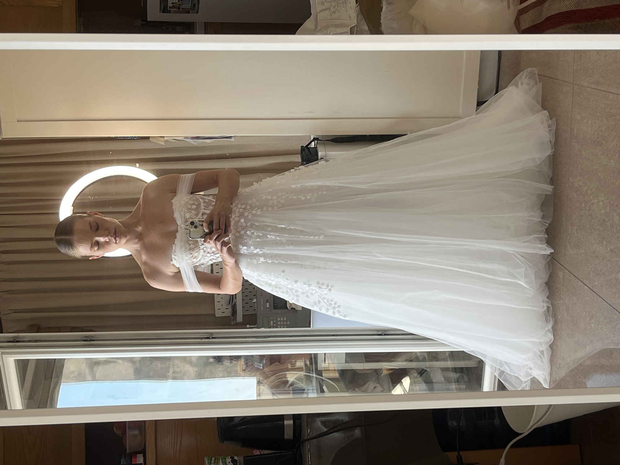 A young woman, likely in her late teens or early twenties, stands in a well-lit room, taking a selfie in a full-length mirror. She is wearing an off-the-shoulder white wedding dress with delicate floral appliques and a voluminous tulle skirt. The dress has a sweetheart neckline and sheer off-the-shoulder straps. Her hair is pulled back in a sleek bun. The background of the image is a reflection in the mirror, revealing a room with beige curtains, a circular ring light, and some kitchen appliances to the left, including a black kettle and a white bucket. A sewing machine is visible on a shelf behind her. The flooring is tiled. The environment suggests an indoor setting, possibly a home or a bridal boutique. No specific landmarks indicating Budva, Montenegro, are identifiable in the image.