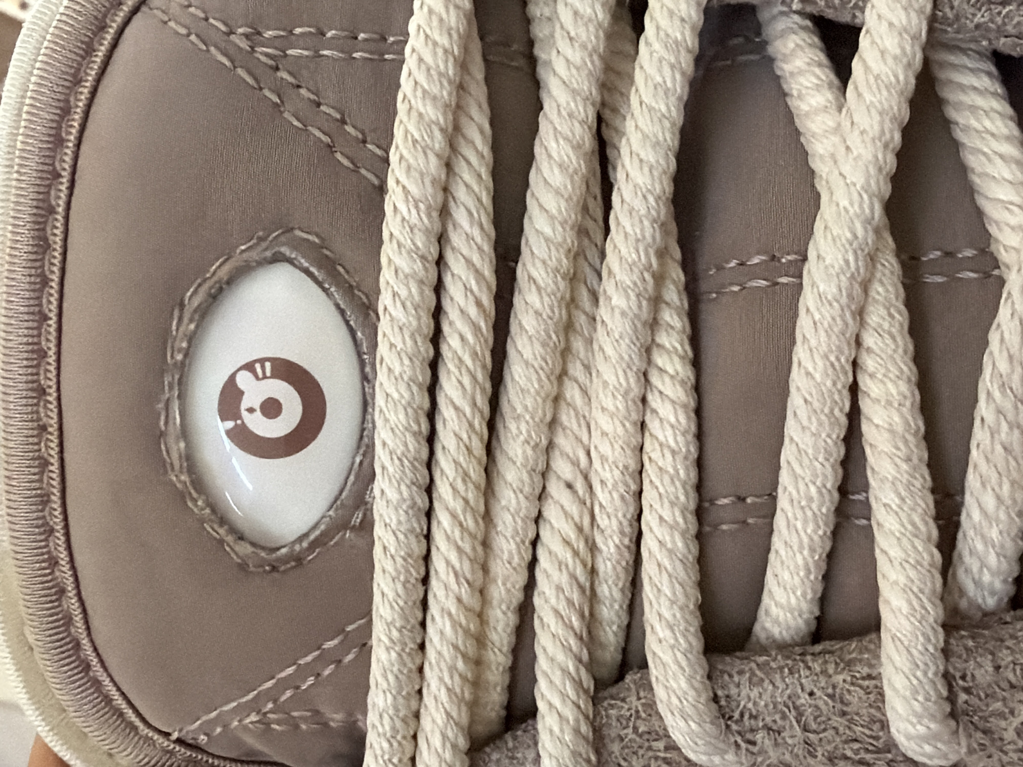The image shows a close-up of a shoe, likely a sneaker, featuring thick, beige, rope-style laces. The shoe itself is a muted brown color, with visible stitching detail. A circular, white badge or emblem with a brown cartoon bear-like face is embedded in the shoe's side panel. The surrounding environment is out of focus, but the image was taken in San Andrés Cholula, Mexico. There are no people visible in the frame, and the primary subject is the shoe itself.