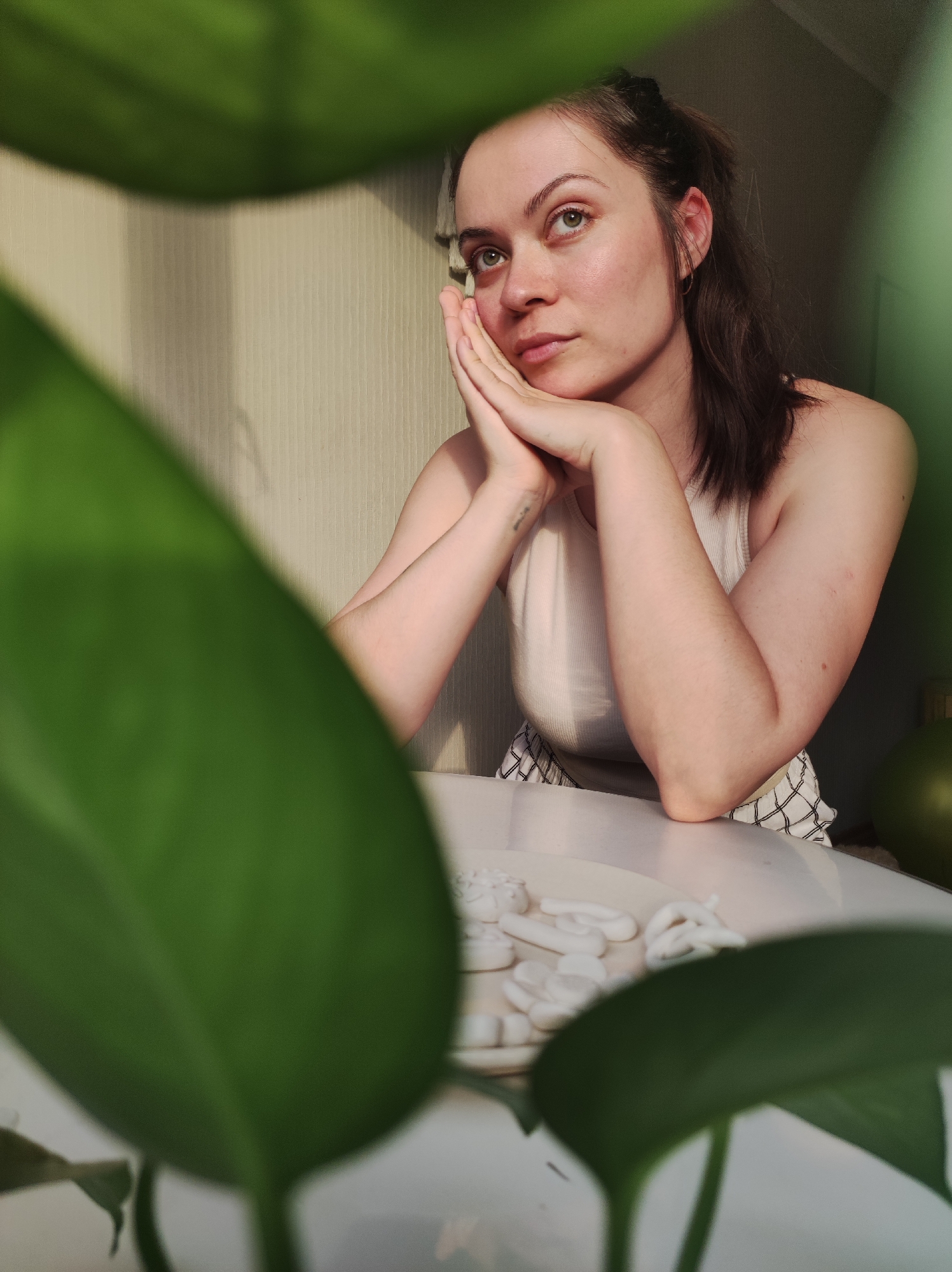 A young woman is sitting at a table with her hands clasped and resting under her chin. She is looking upward and to the left with a thoughtful expression. She has brown hair pulled back, and is wearing a white tank top. In front of her on the white table is a plate with white objects that appear to be decorative. The scene is framed by large green leaves in the foreground, suggesting a plant is nearby. The wall behind her is light beige with a subtle vertical pattern. A portion of a green spherical object is visible to the right of the woman. The location is Chaikovsky, Russia.