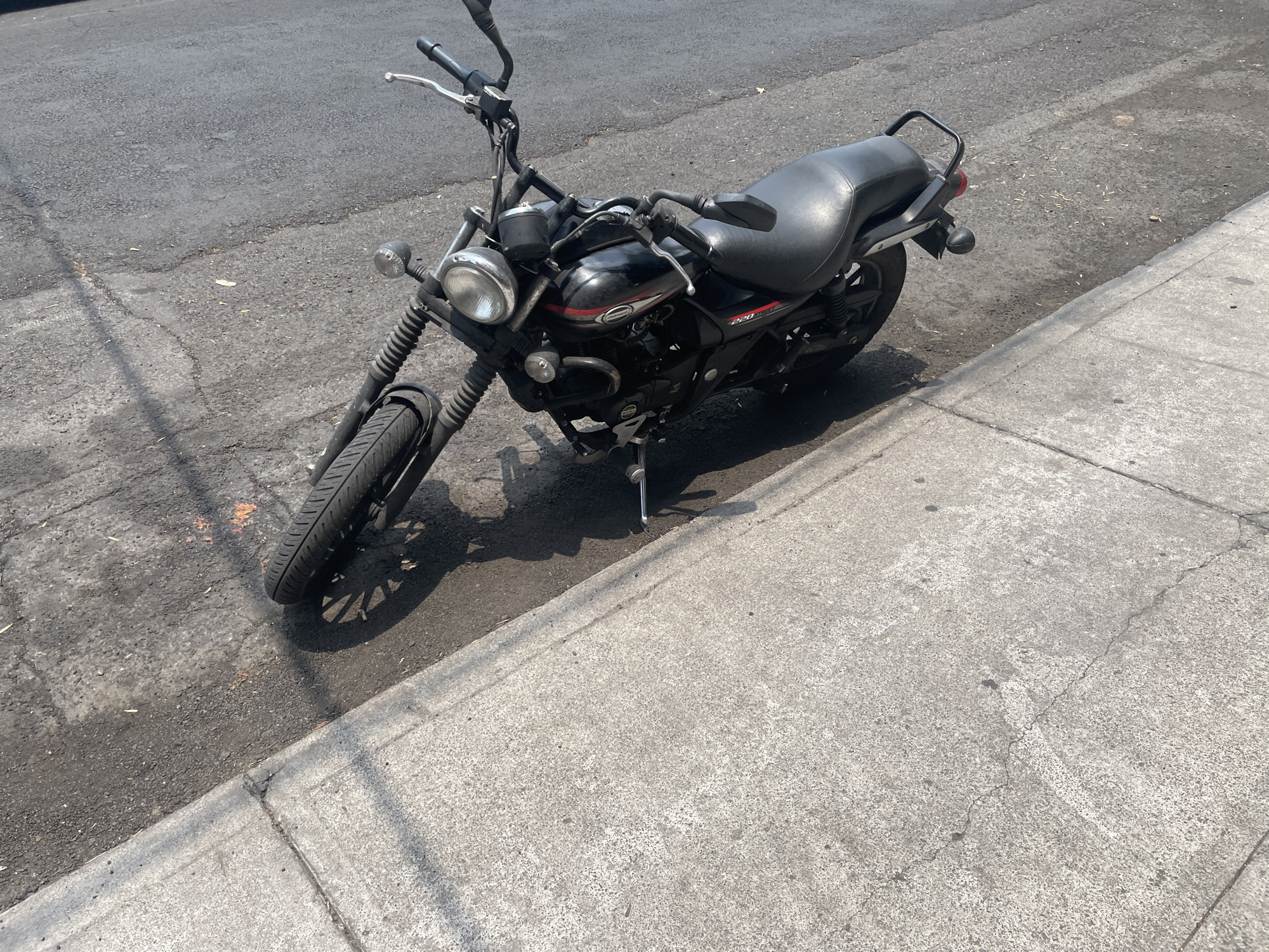 The image shows a black Bajaj Avenger 220 Street motorcycle parked on a street in Mexico City, Mexico. The motorcycle is parked on the asphalt near the concrete sidewalk. The scene is lit by sunlight, casting a shadow of the motorcycle on the asphalt. The street is asphalt, and the sidewalk is concrete. The motorcycle has a black seat, black fuel tank with red and silver details, a round headlight, and black handlebars.