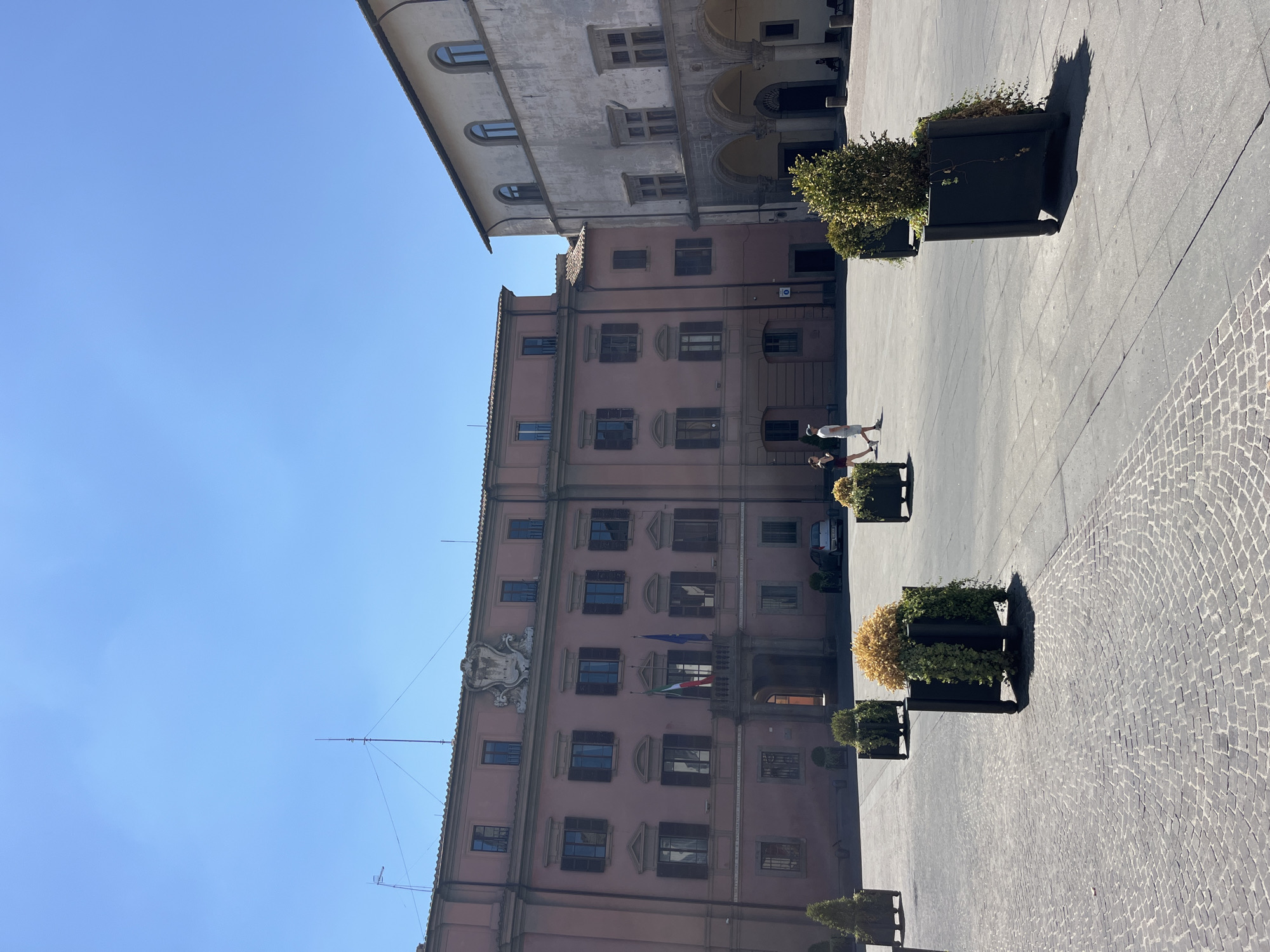 The image depicts a bright, sunny day in a European town square. Two prominent buildings dominate the scene. The building on the left is a large, pink structure with multiple windows and a coat of arms above the central entrance. Two flags, one Italian and one likely European Union, fly in front of the entrance. The building on the right is a lighter-colored stone structure with arched walkways on the ground floor and arched windows on the upper floors.
The square itself is paved with cobblestones and large square tiles. Several large, dark planters containing green bushes and some dry, yellow foliage are strategically placed throughout the square. In the foreground, a few people are walking, one couple is walking away from the camera and another person is walking towards the camera. A silver car is parked near the pink building. The sky is clear and blue.
The location is Piazza del Comune in Viterbo, Italy.