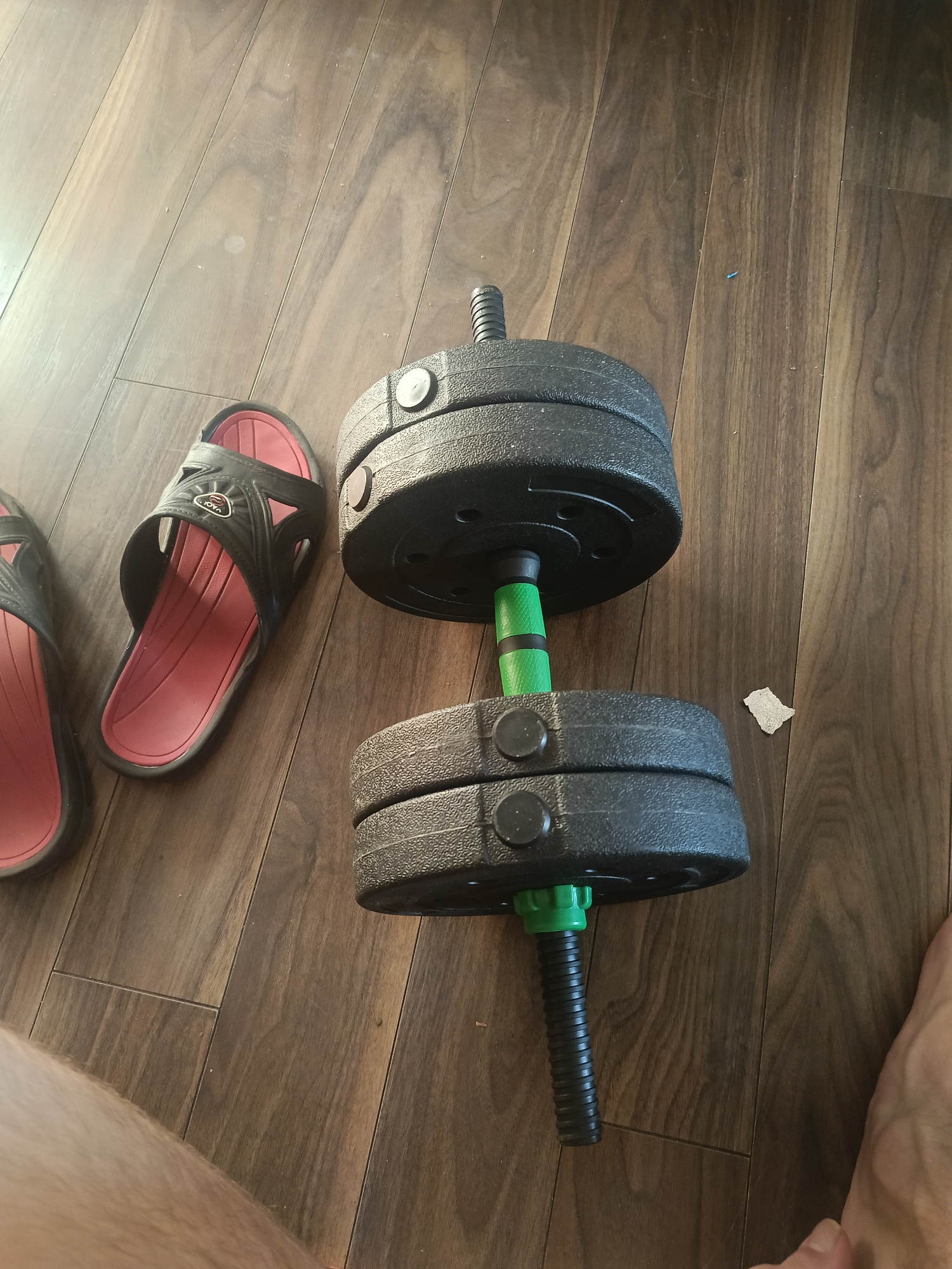 An adjustable dumbbell and two black and red slides are arranged on a wooden floor. The dumbbell has two black weight plates on each side of a green handle. The slides are positioned to the left of the dumbbell, with their soles facing upwards. The scene is likely indoors, possibly in a home or gym setting, with the wooden floor suggesting a residential or exercise facility. The lighting indicates a well-lit environment, possibly natural light from a window or overhead artificial lighting. The image provides no information about the city of Grodzisk Mazowiecki, Poland, as it is a visual description of objects and their placement.